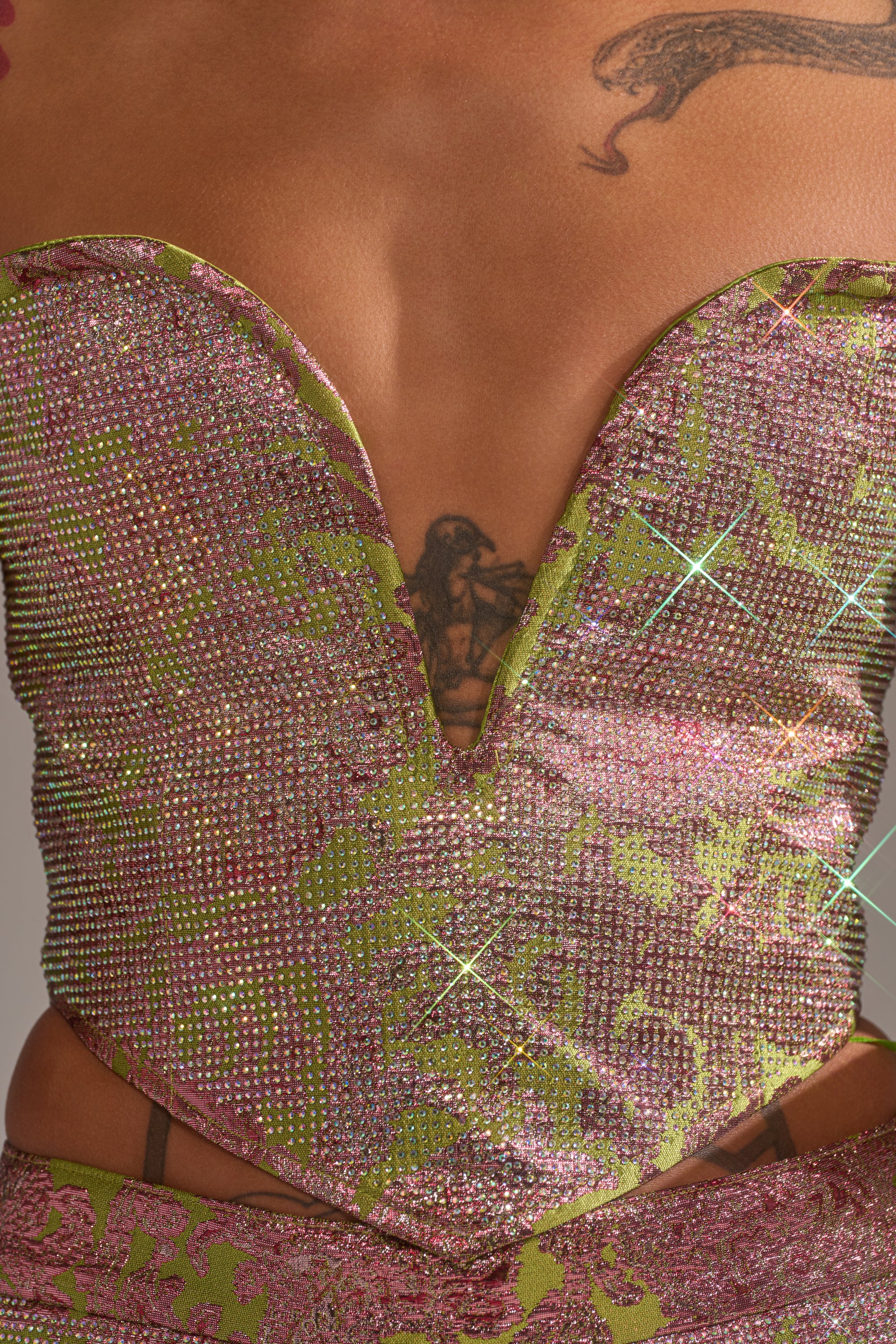 A close-up of a person wearing the TIME AND TIME AGAIN RHINESTONE CORSET by AKIRA Label, featuring pink and green rhinestones, a sparkling strapless design, deep V neckline, and a visible chest tattoo above the neckline. 7