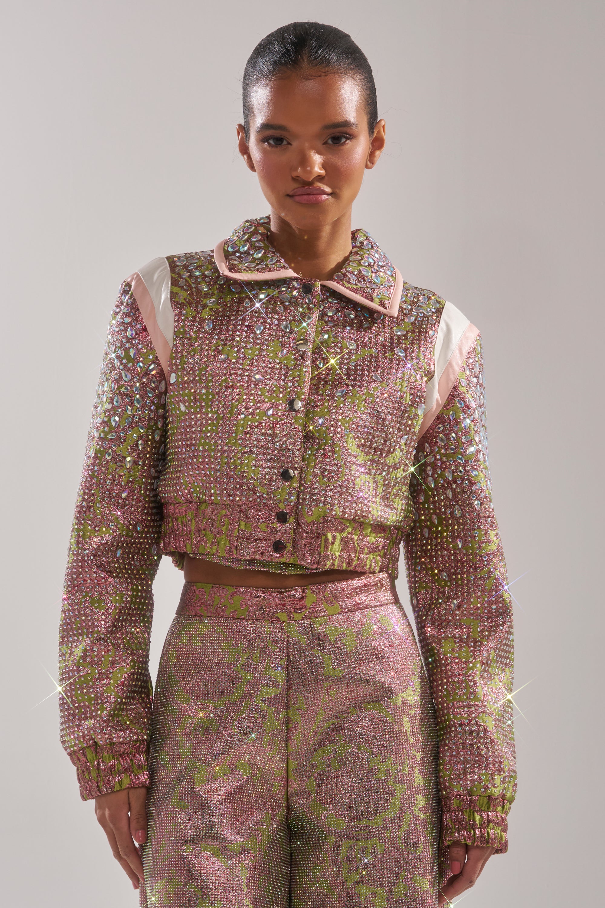 A person wearing the TIME AND TIME AGAIN RHINESTONE BOMBER, a sparkling pink and gold sequined jacket with matching high-waisted pants, stands against a plain light background. 0