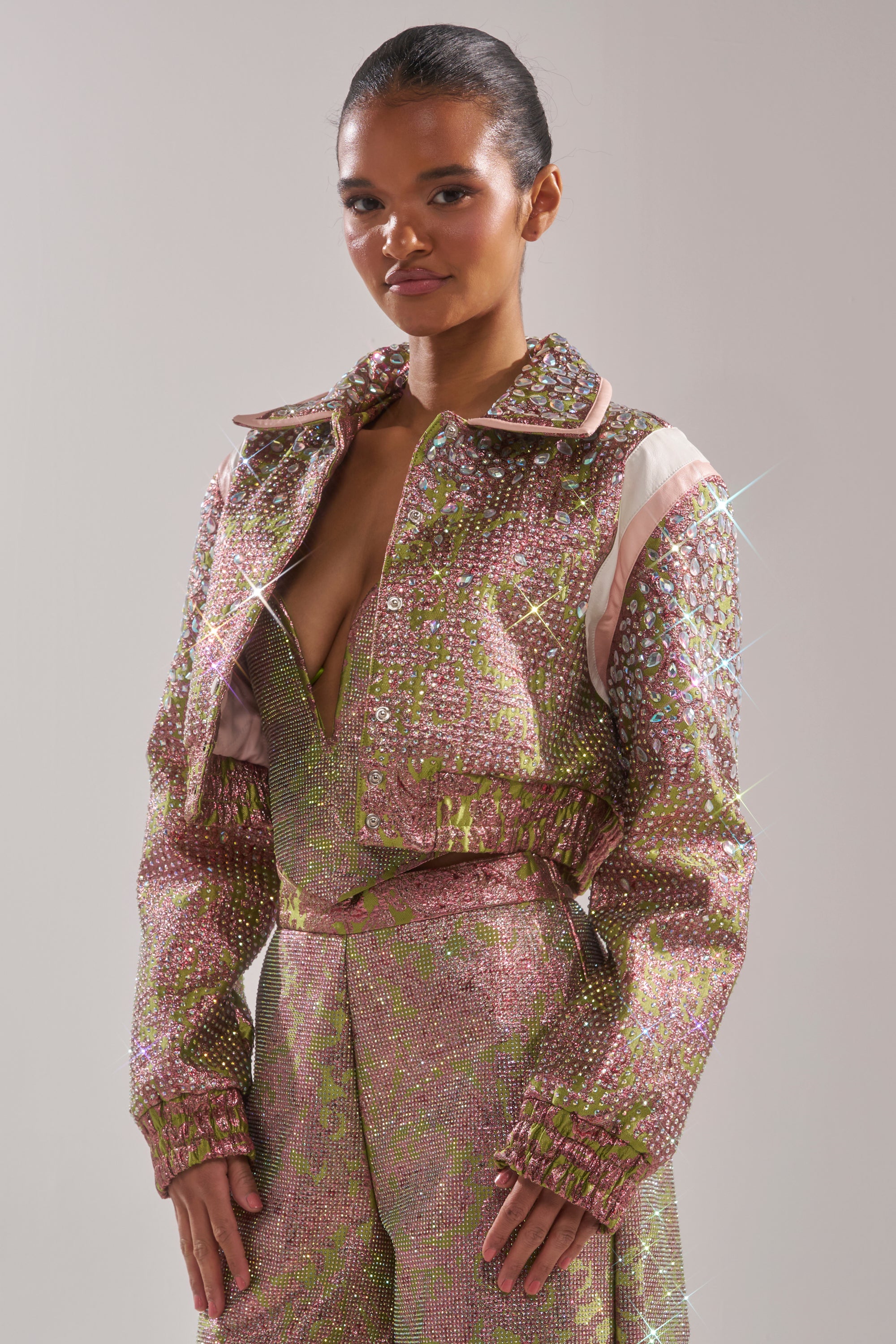 A woman stands against a plain background wearing the TIME AND TIME AGAIN RHINESTONE BOMBER, a shimmering tapestry-inspired jacket with sparkling details and a deep neckline, her hair pulled back in a sleek style. 3