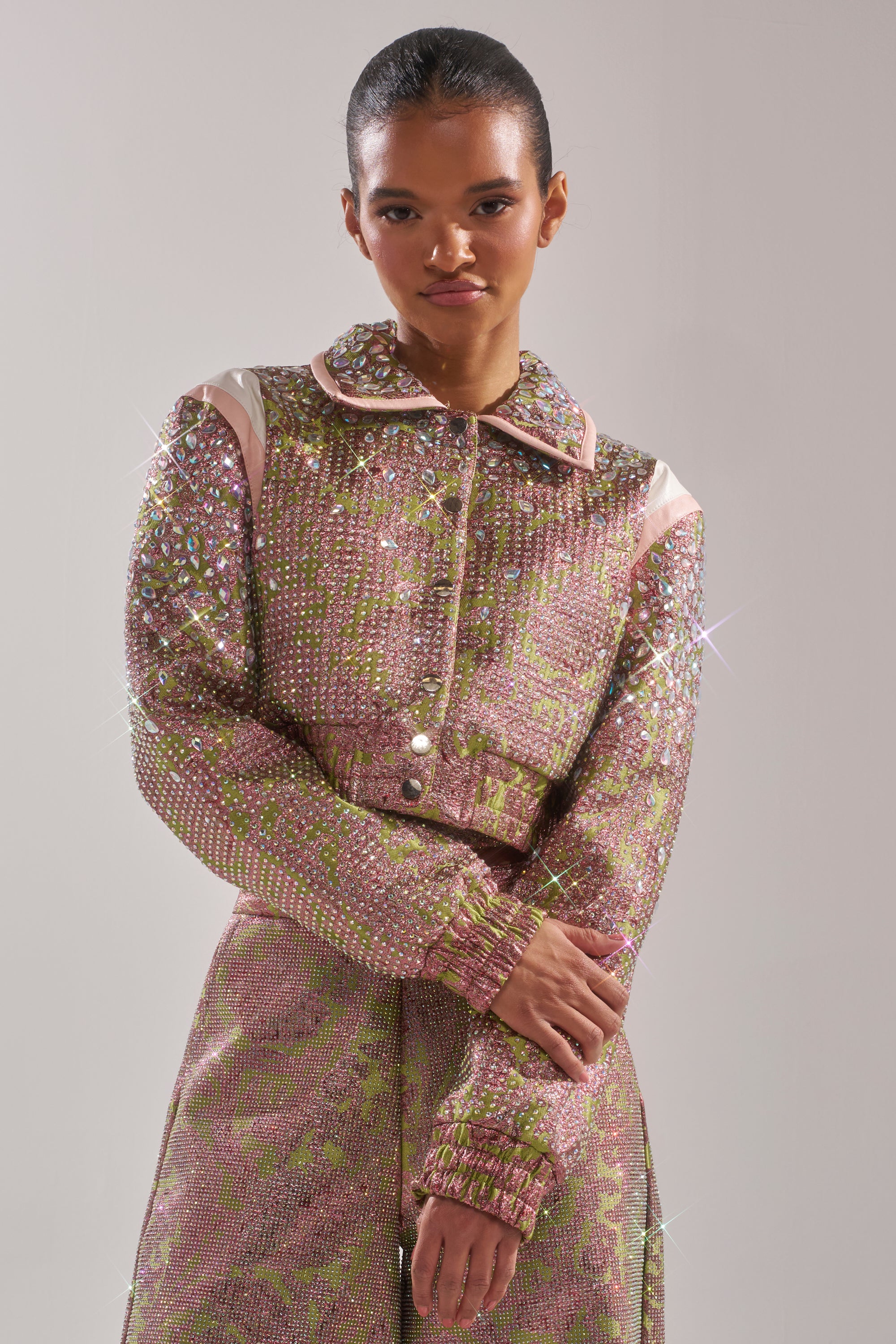 A woman with slicked-back hair crosses her arms while wearing the TIME AND TIME AGAIN RHINESTONE BOMBER and matching skirt, featuring sparkling pink and green rhinestones, in front of a plain light background. 4