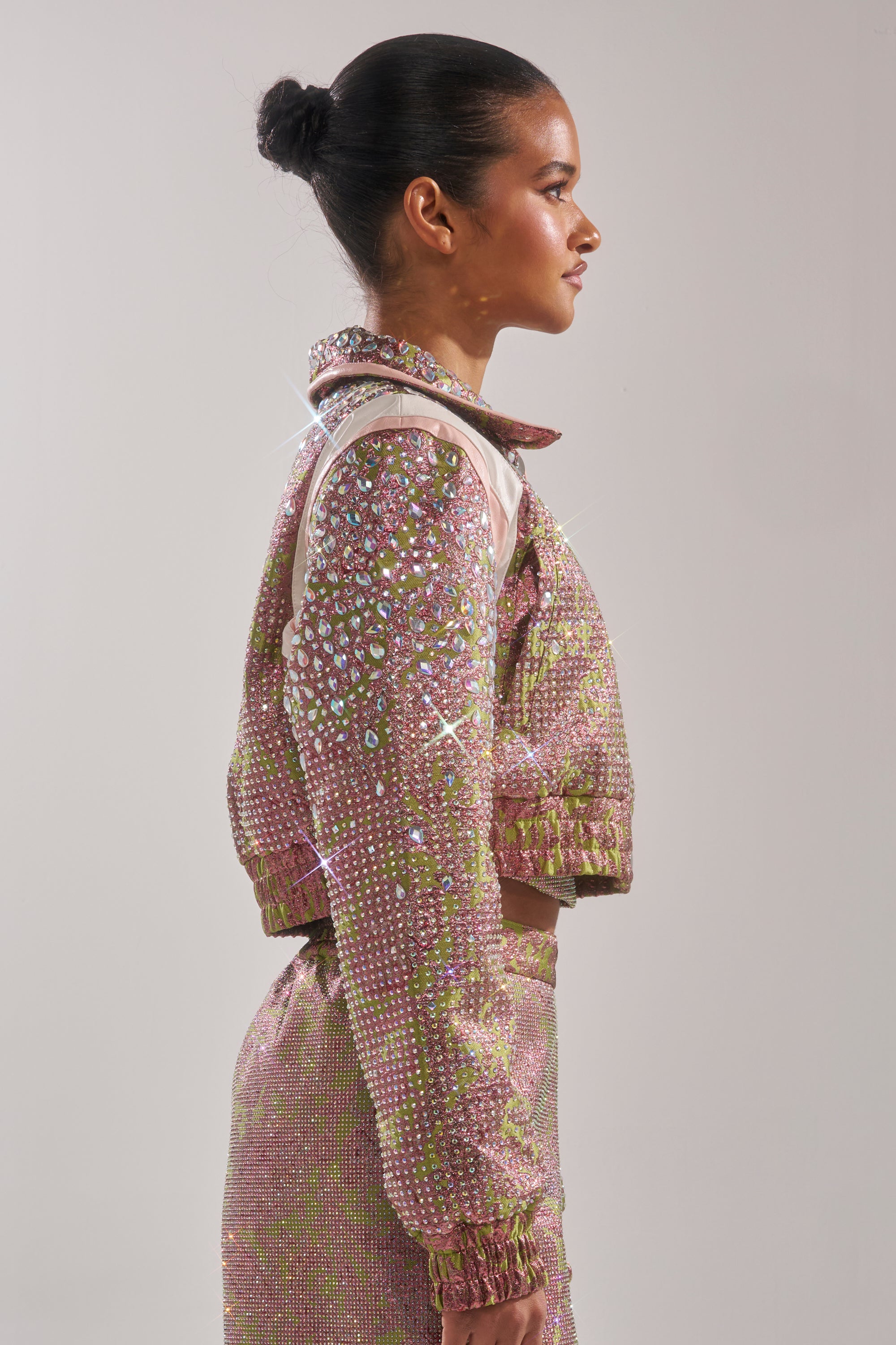 A woman with a neat bun stands in profile against a plain light background, wearing the sparkling pink and green TIME AND TIME AGAIN RHINESTONE BOMBER and matching skirt, both embellished with shiny rhinestones. 5