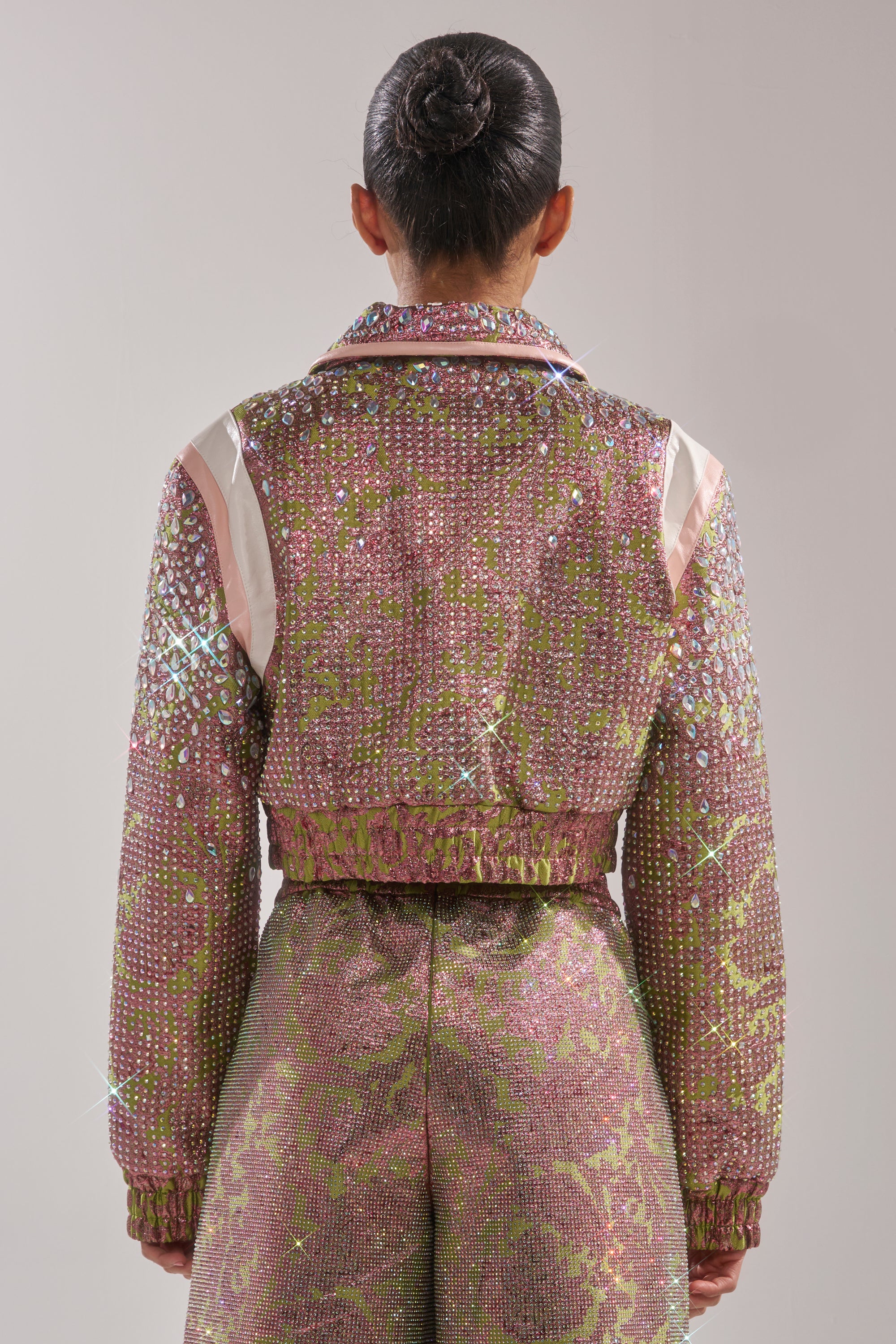 A person with dark hair in a bun is shown from behind, wearing the TIME AND TIME AGAIN RHINESTONE BOMBER covered in sparkling pink and green rhinestone patterns and crystal embellishments. 6