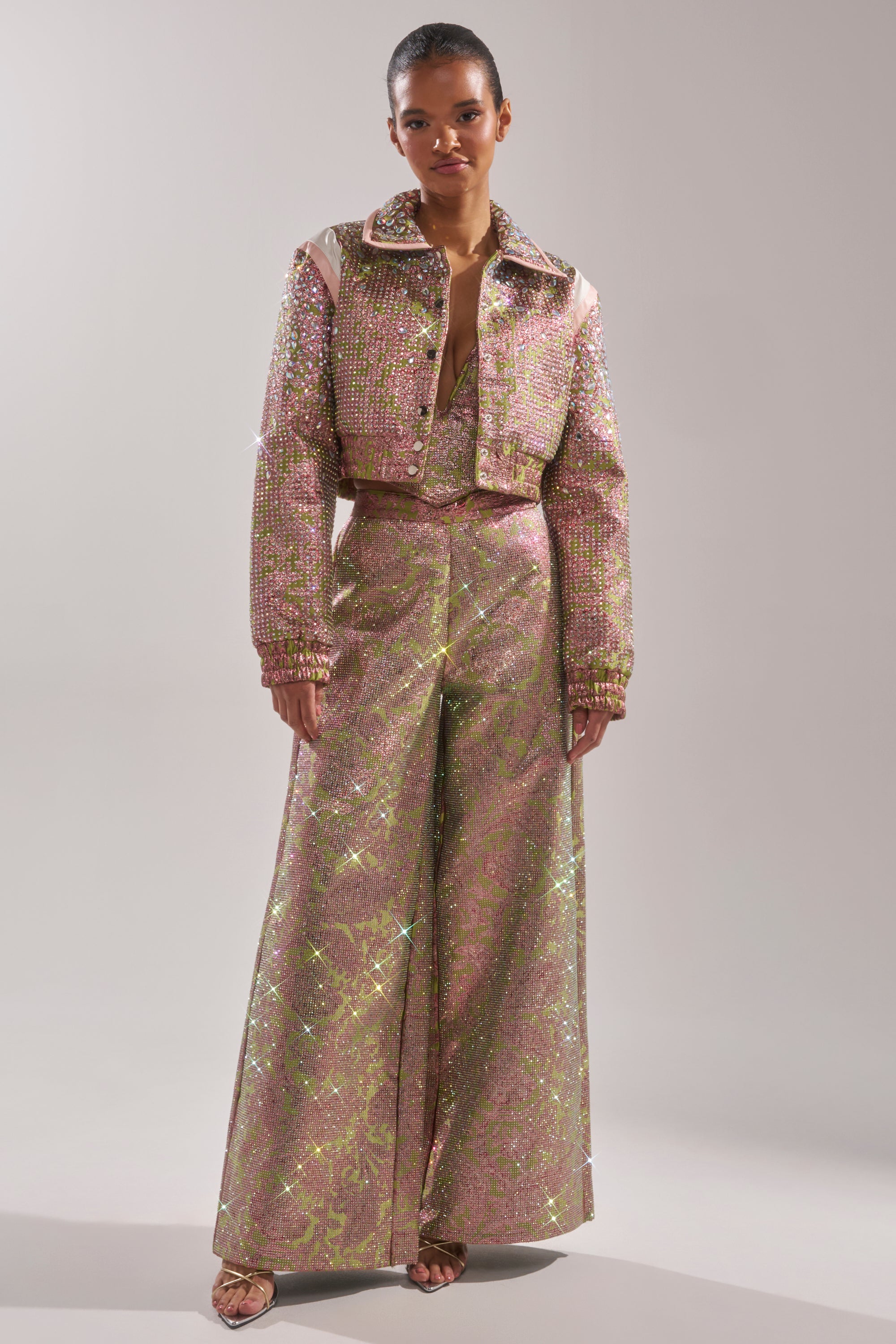 A person wears the TIME AND TIME AGAIN RHINESTONE BOMBER, a sparkling light pink crystal-embellished cropped jacket with matching high-waisted wide-leg pants and open-toe heels, reflecting light for a shimmering effect. 7