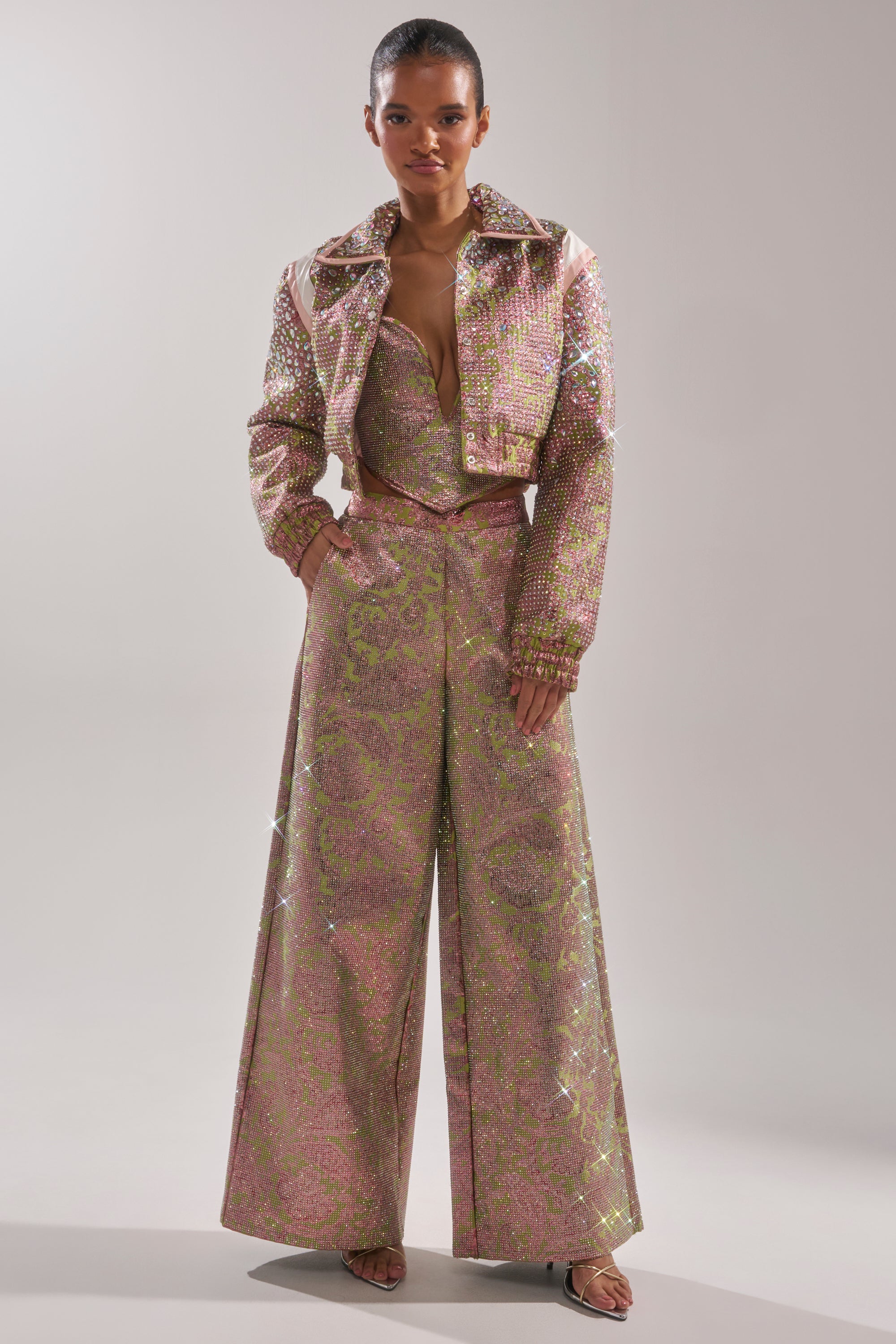 A woman wears the TIME AND TIME AGAIN RHINESTONE BOMBER, paired with a sparkling pink and green wide-leg pantsuit featuring a deep V-neck bustier, her hair tied back and open-toe sandals, posing against a plain background. 8
