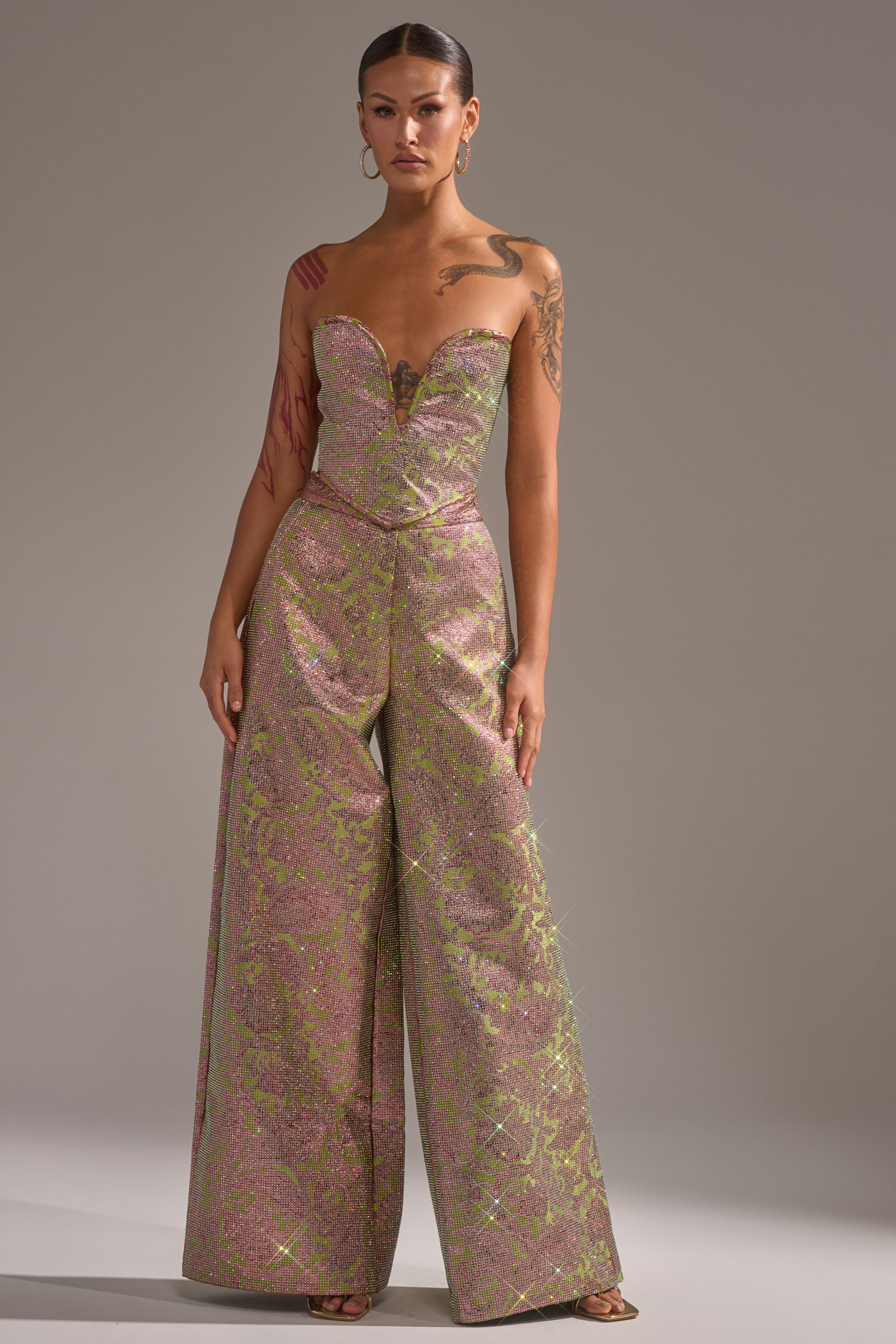 A woman wears the Time and Time Again Rhinestone Trouser, featuring a metallic pink and green floral brocade, wide-leg silhouette, and deep sweetheart neckline. She styles it with slicked-back hair, hoop earrings, and shows off her arm tattoos. 1