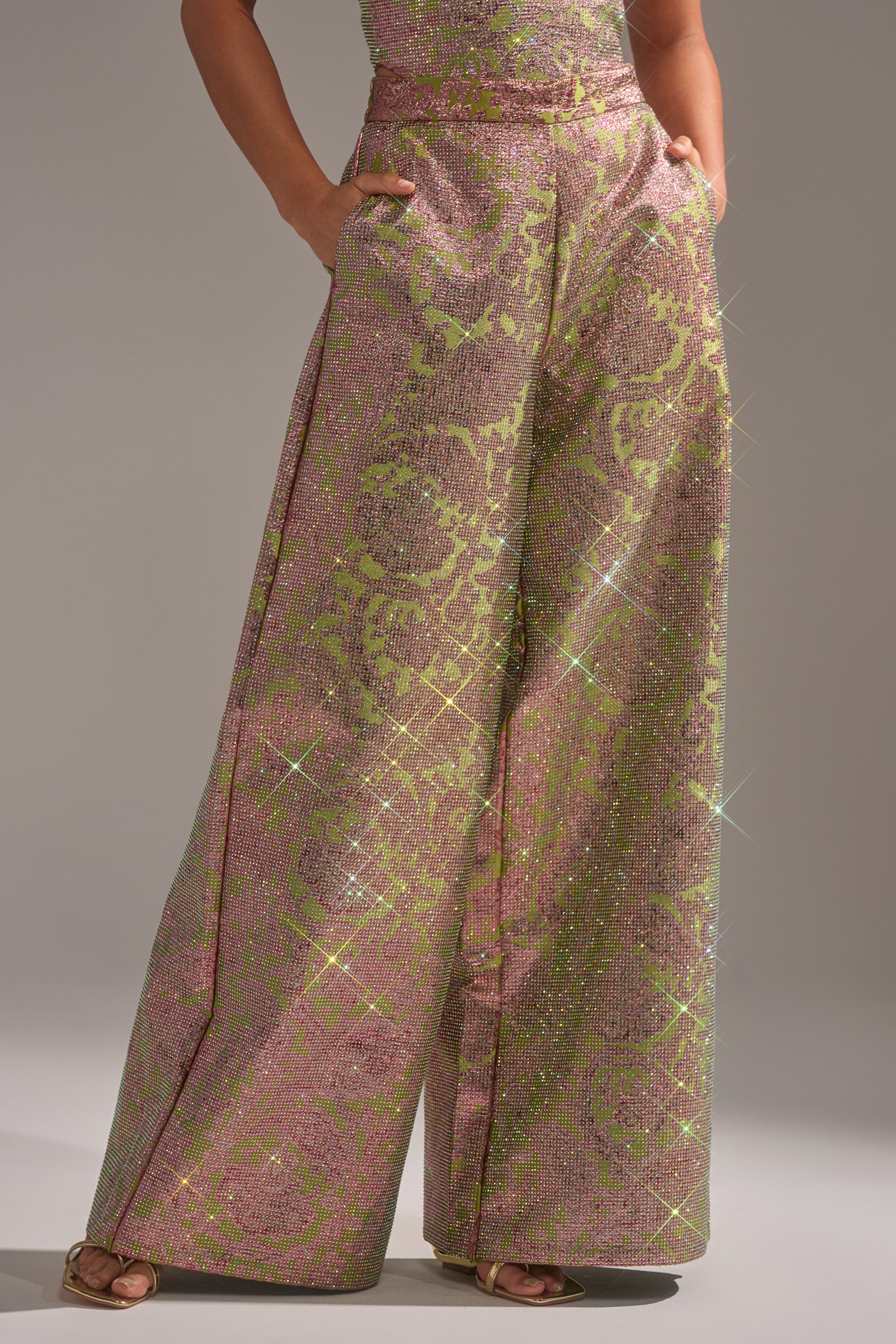 Someone stands with hands in pockets, slightly turned to the side, wearing the TIME AND TIME AGAIN RHINESTONE TROUSER—high-waisted wide-leg pants in sparkling pink and green—with a plain background. 0