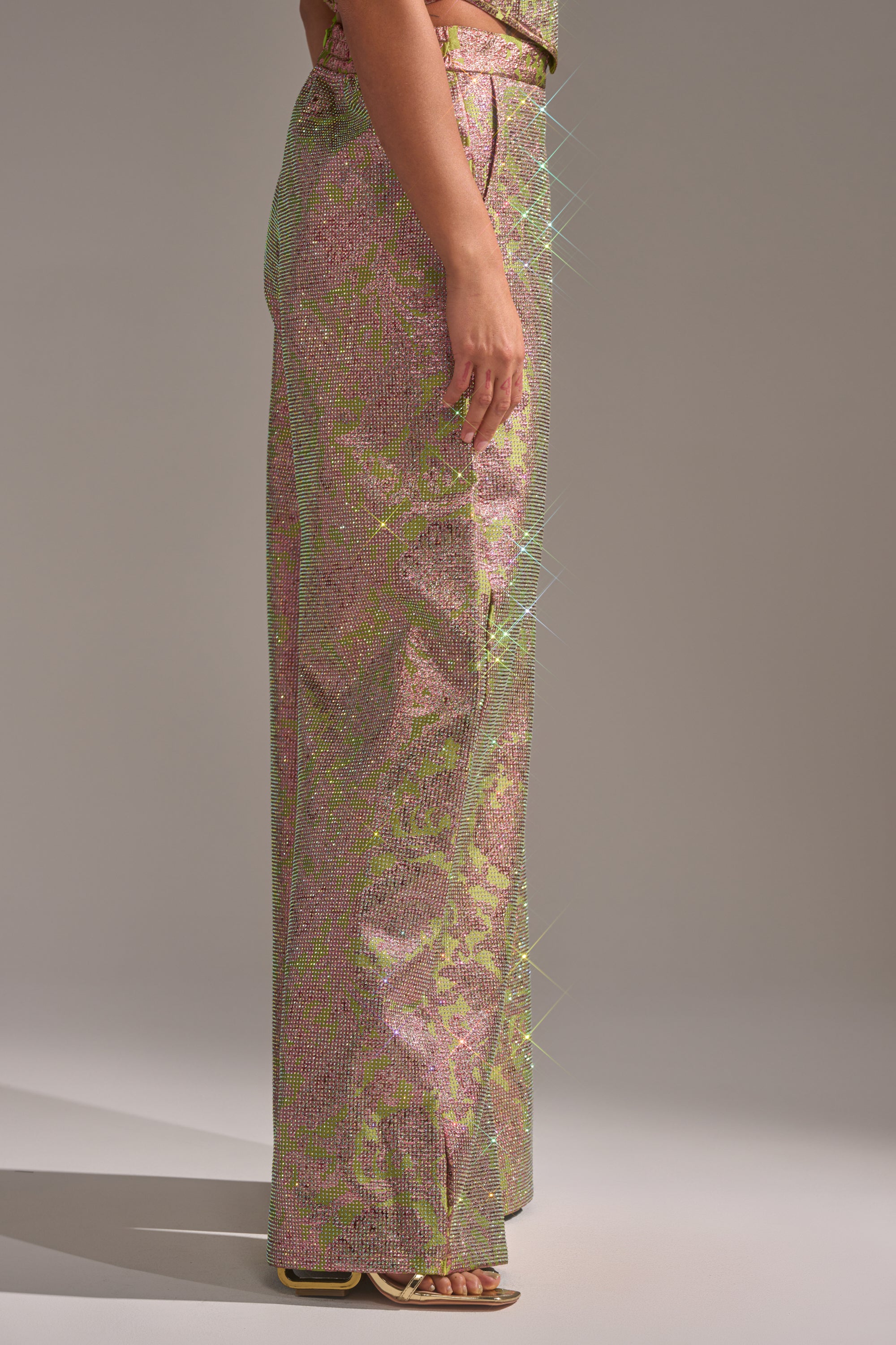 A person stands sideways against a plain light gray background, wearing the TIME AND TIME AGAIN RHINESTONE TROUSER—high-waisted, wide-leg pants with sparkly pink and green sequins—paired with open-toe sandals. 2