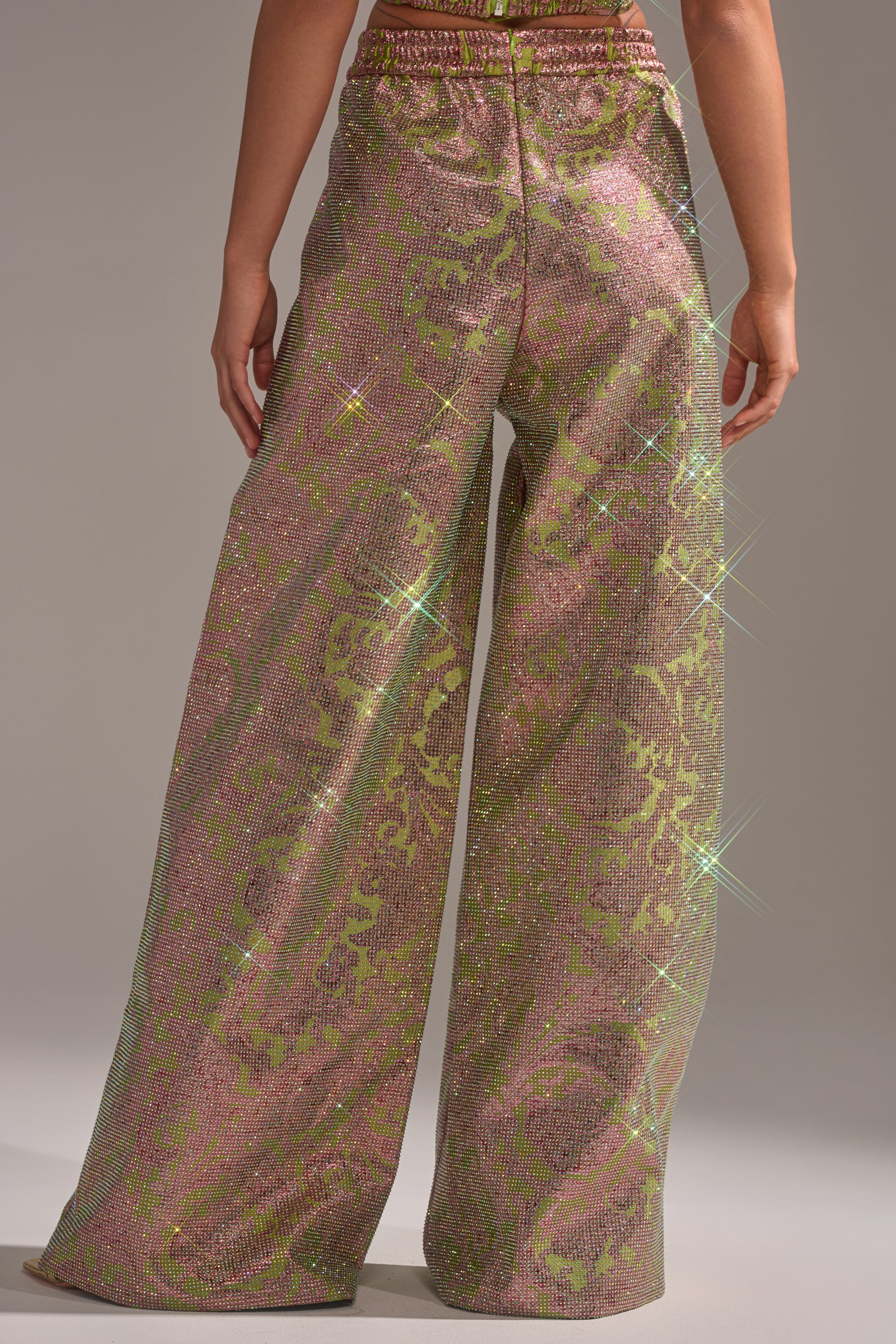 A person shown from the back wearing the TIME AND TIME AGAIN RHINESTONE TROUSER—high-waisted, wide-leg pants featuring a shimmering pink and green floral brocade with sparkly rhinestone accents. 3