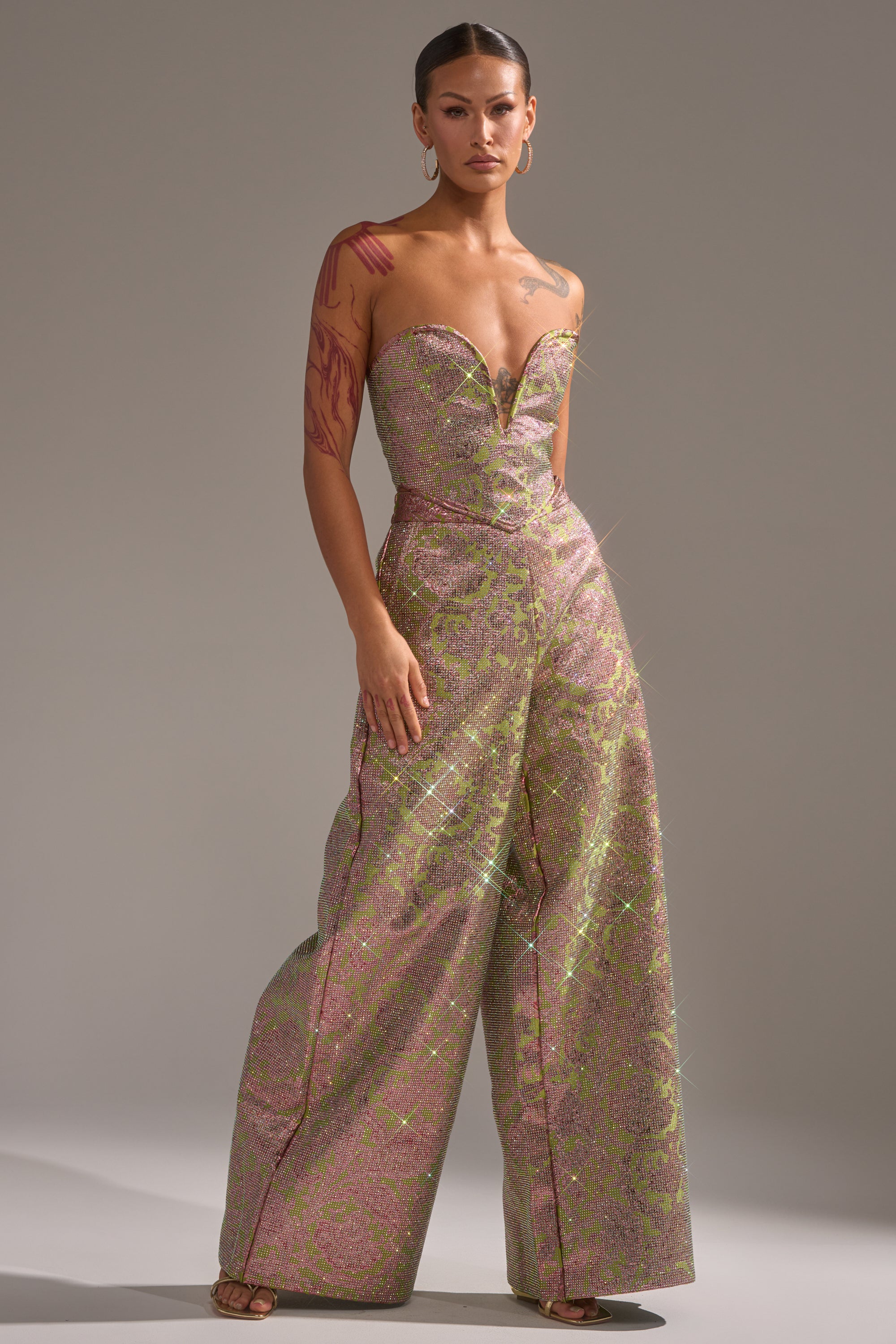 A woman stands confidently in the TIME AND TIME AGAIN RHINESTONE TROUSER, a strapless, sparkling wide-leg jumpsuit with a metallic pink and green floral pattern. She wears hoop earrings, has slicked-back hair, and tattoos on her right arm. 5