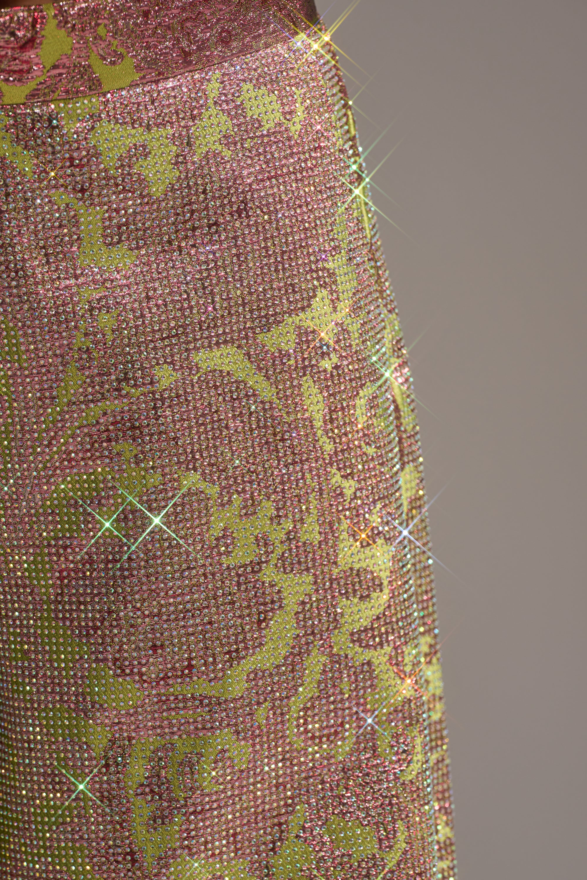Close-up of the TIME AND TIME AGAIN RHINESTONE TROUSER's shimmering pink and gold floral brocade fabric, adorned with sparkling rhinestones that reflect light. The ornate pattern stands out against a softly blurred neutral background. 6