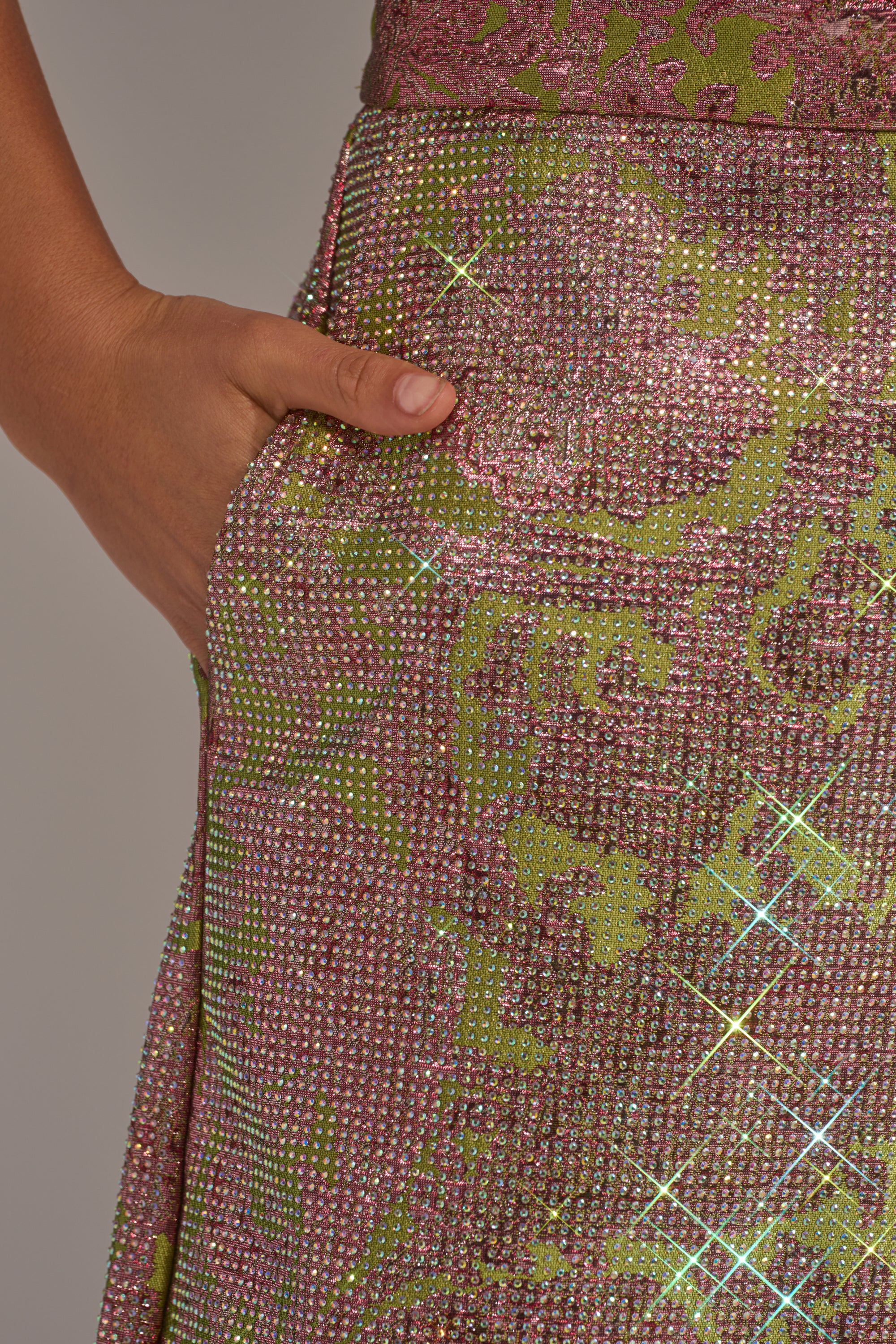 A close-up of a hand resting in the pocket of the TIME AND TIME AGAIN RHINESTONE TROUSER, featuring sparkling multicolored floral brocade with green and pink patterns and glittering rhinestone accents. 7