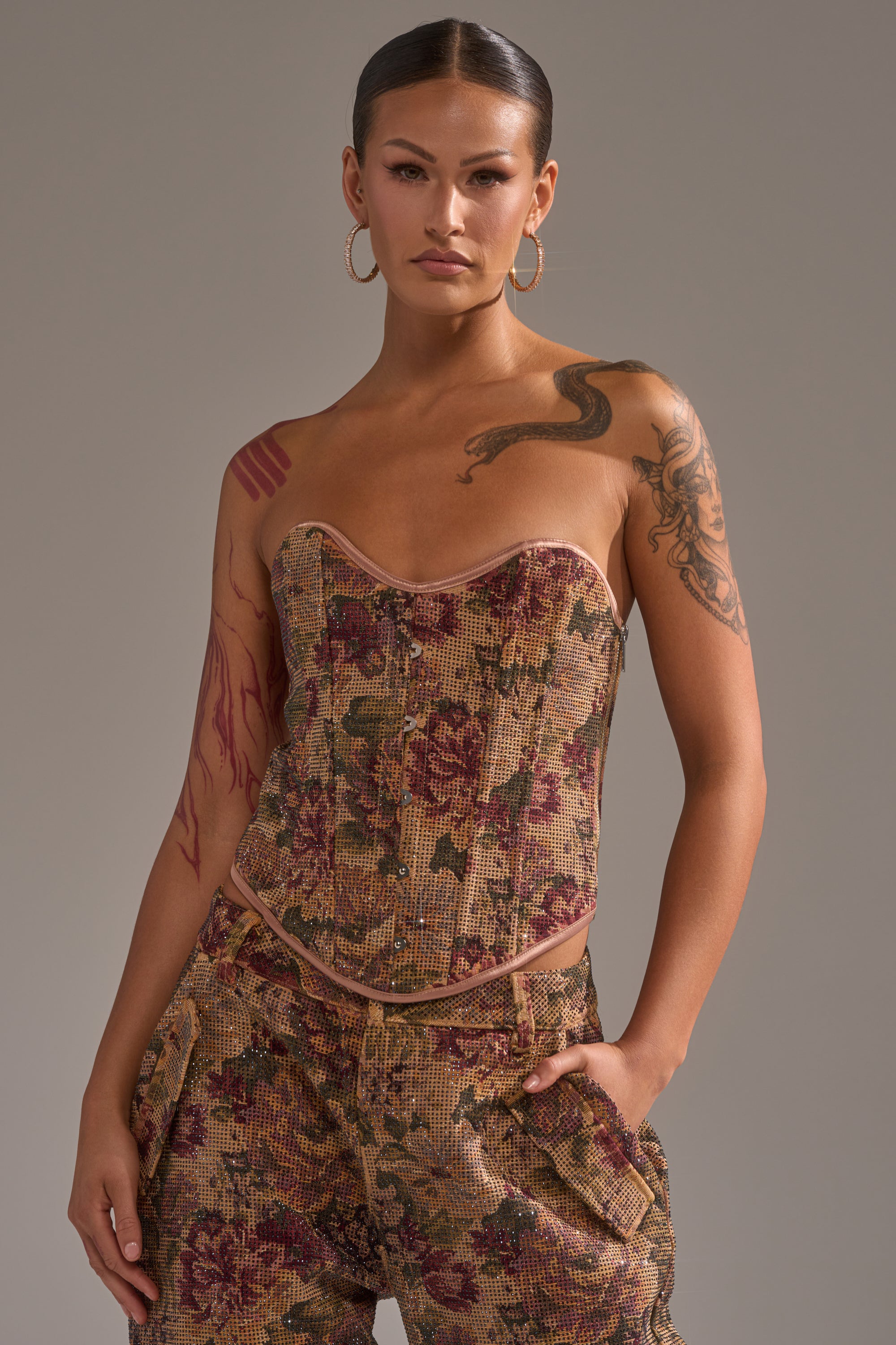 A tattooed individual wears the GLAM SQUAD RHINESTONE BROCADE CORSET with matching pants, hands in pockets, posing confidently against a plain gray background.