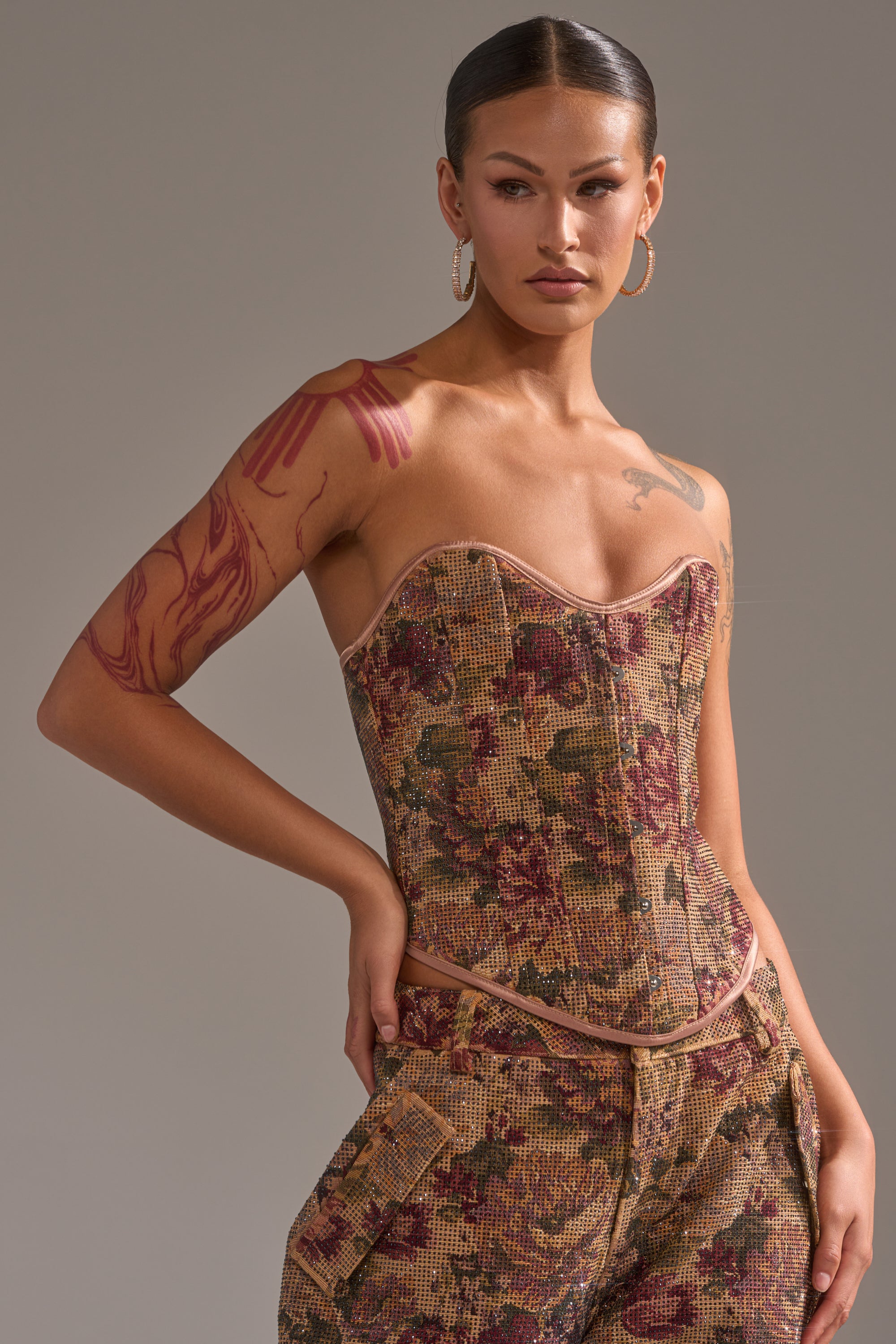 A woman poses in the GLAM SQUAD RHINESTONE BROCADE CORSET with matching pants, featuring a strapless sweetheart neckline and floral jacquard. She has slicked-back hair, large hoop earrings, and visible tattoos against a light gray background.