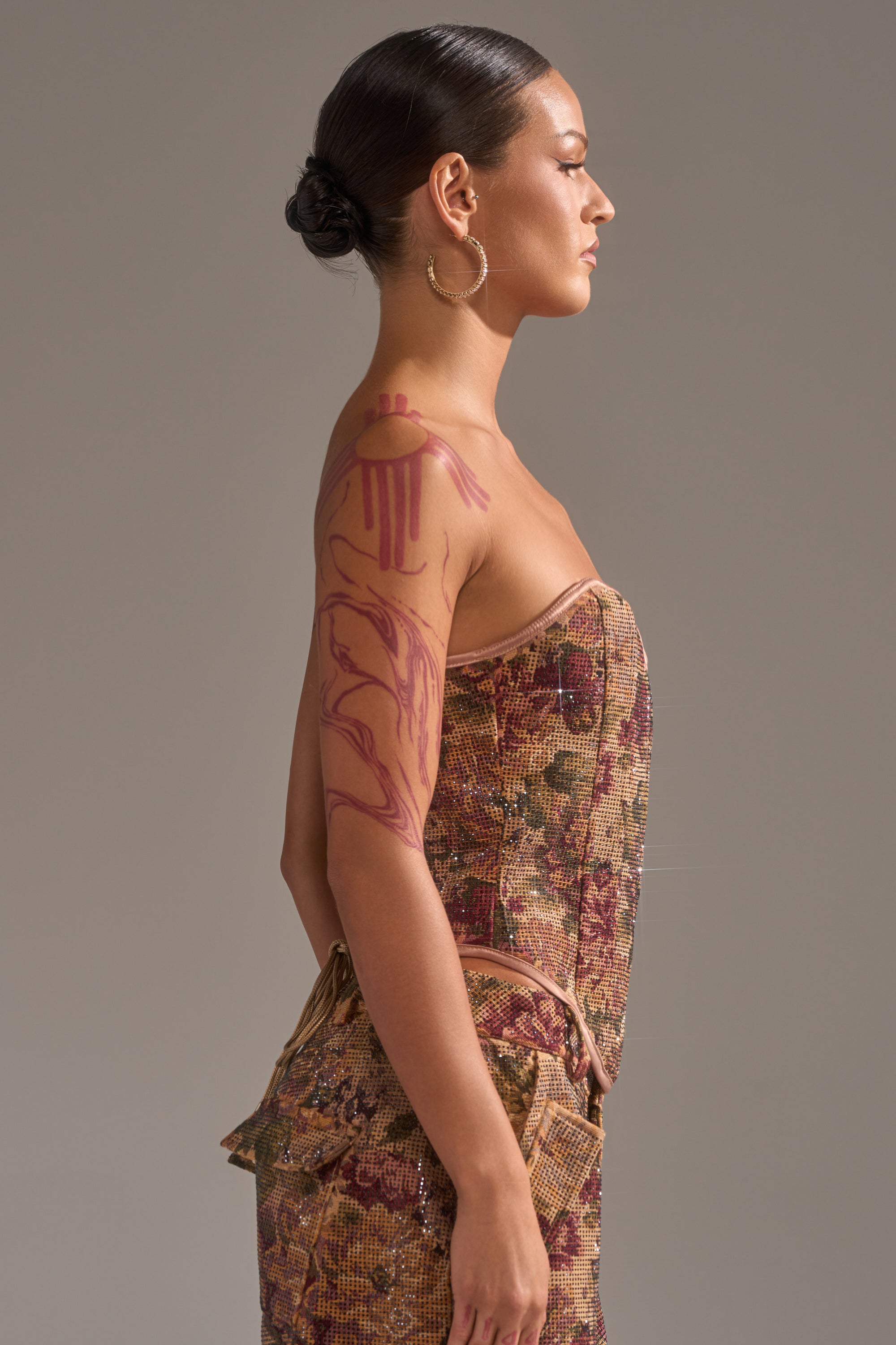 A woman with a sleek bun wears the GLAM SQUAD RHINESTONE BROCADE CORSET with a sweetheart neckline and floral pattern, holds a matching clutch, and shows off red abstract arm art while standing in profile against a plain backdrop.