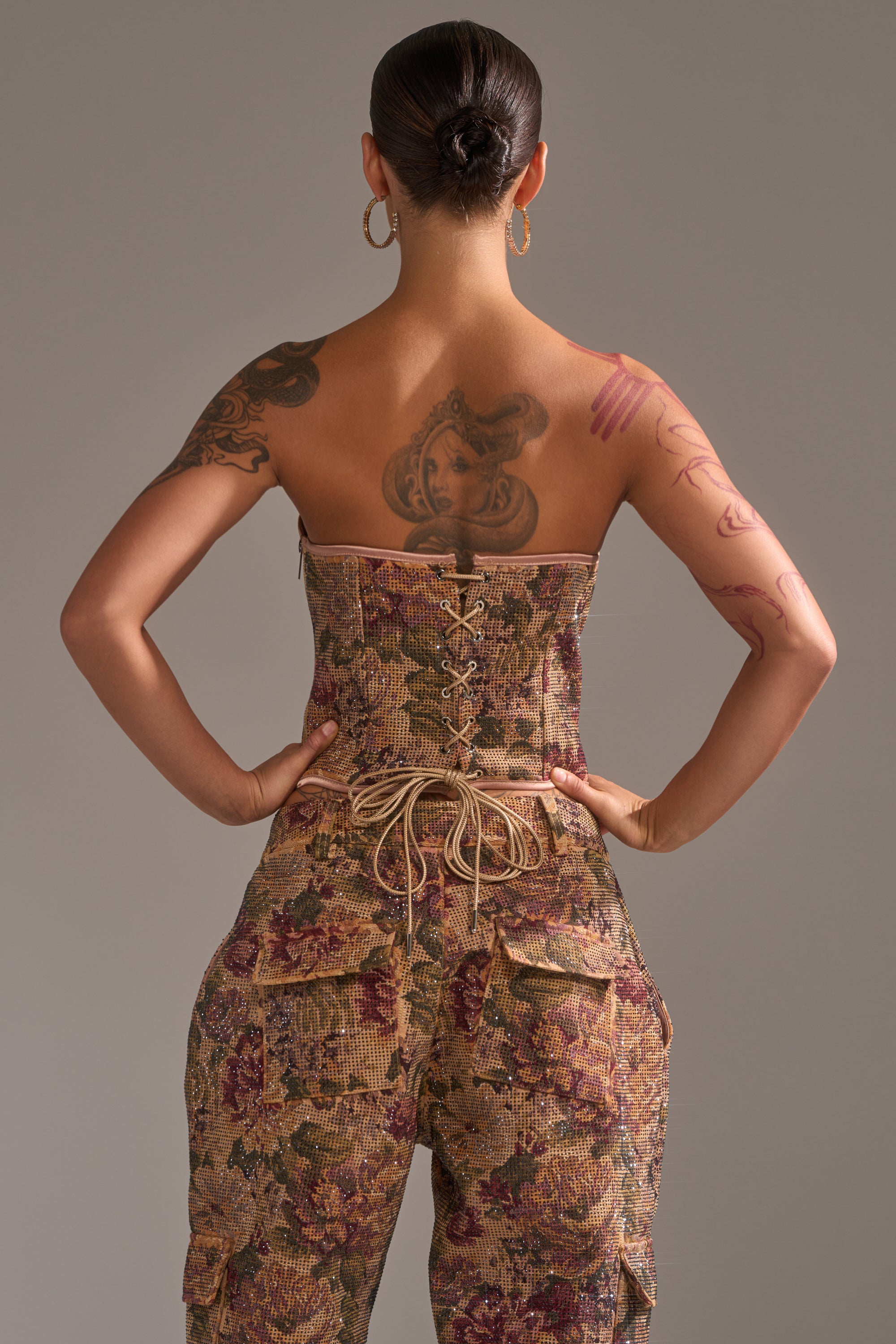 A tattooed person with hoop earrings and a bun stands facing away, hands on hips, wearing the GLAM SQUAD RHINESTONE BROCADE CORSET and matching cargo pants against a plain background.