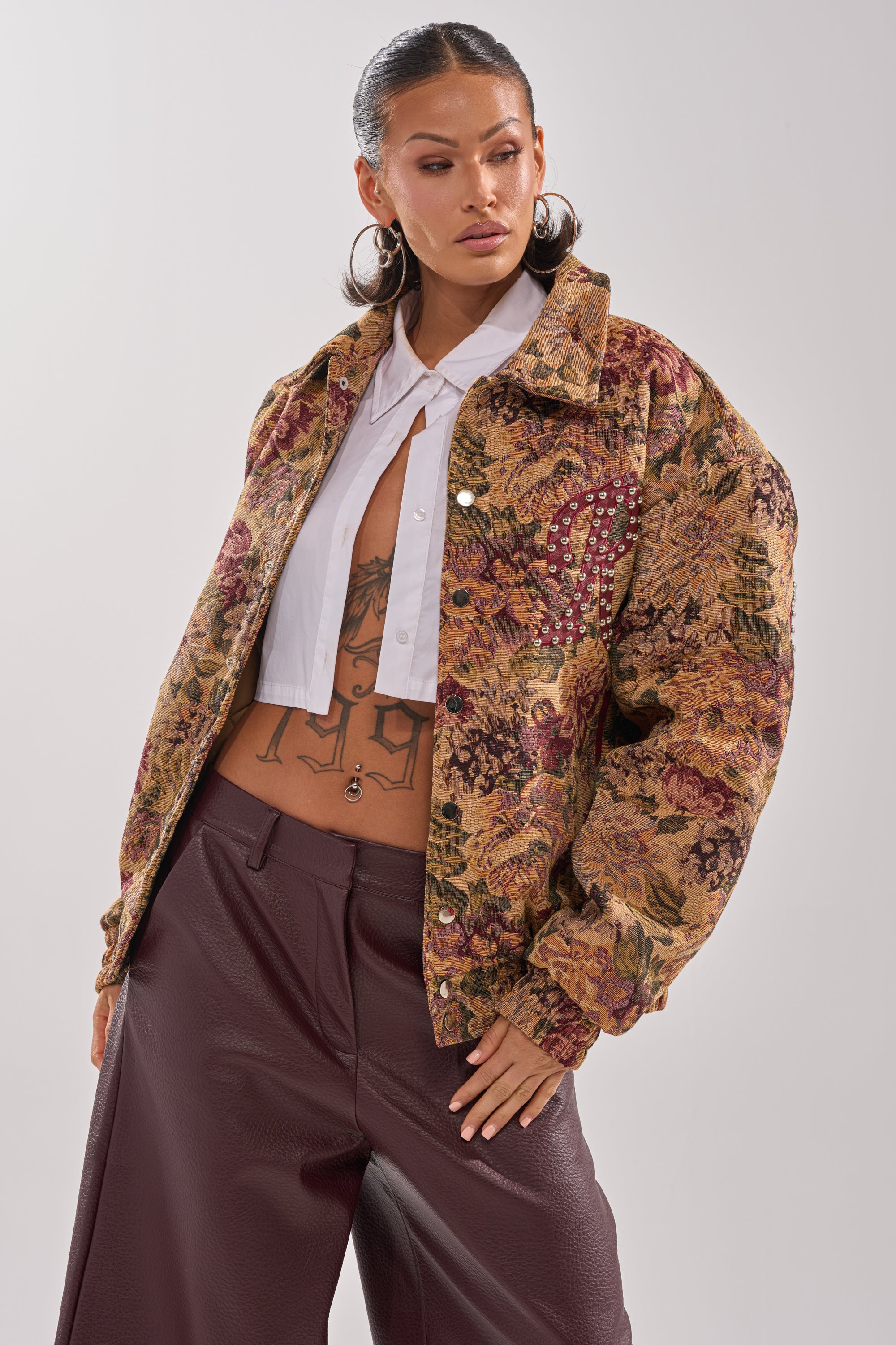A woman confidently poses in the SLIDE ON ME TAPESTRY BOMBER, paired with a white cropped blouse and high-waisted maroon pants. She has slicked-back hair, hoop earrings, and visible torso tattoos. 0