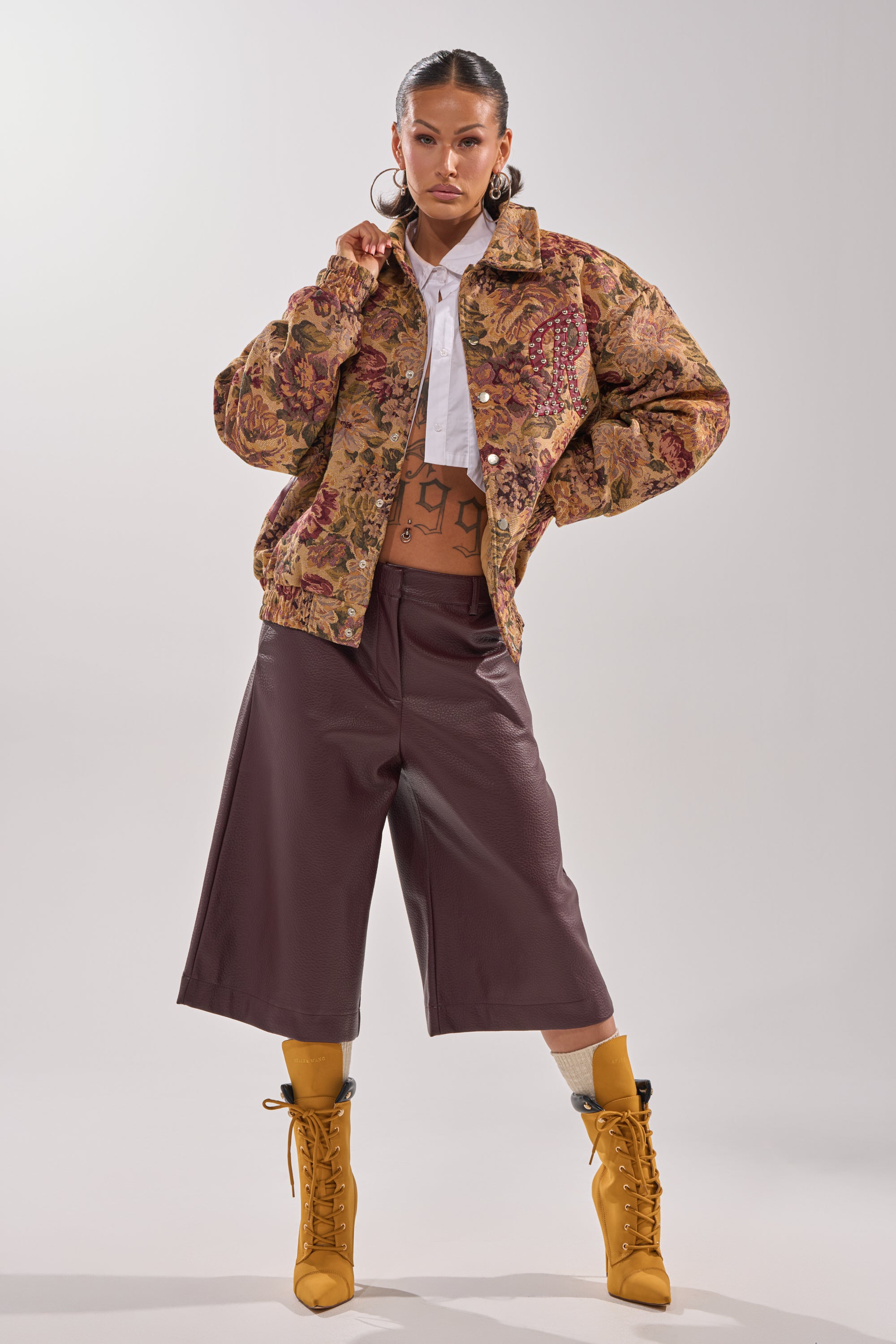 A person exudes confidence in the SLIDE ON ME TAPESTRY BOMBER, styled with a white cropped top, maroon culottes, mustard lace-up boots, bold hoop earrings, and sleek slicked-back hair. 9