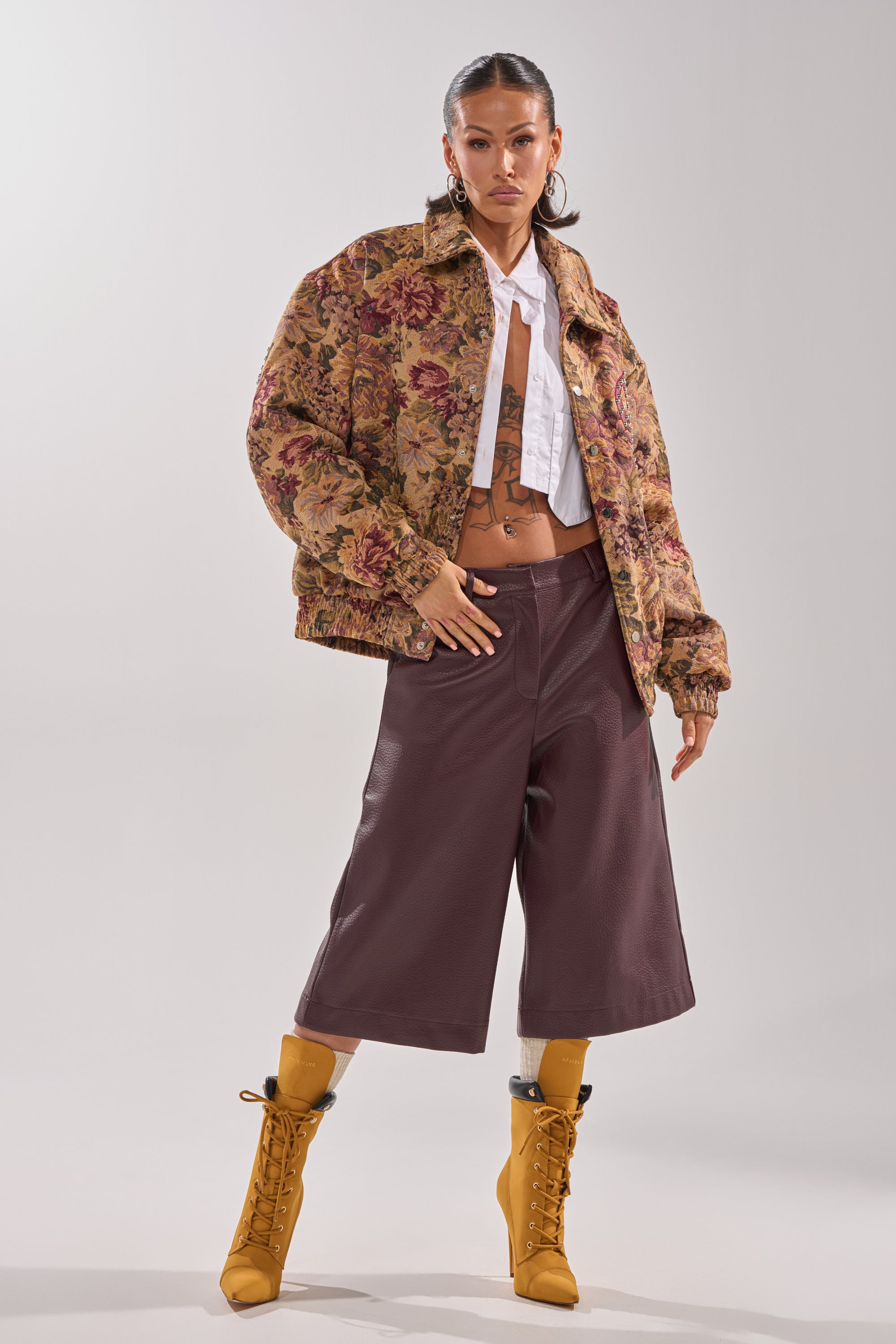A person poses in the SLIDE ON ME TAPESTRY BOMBER, an unbuttoned white shirt, high-waisted brown culottes, and mustard yellow heeled boots against a plain light background. 10