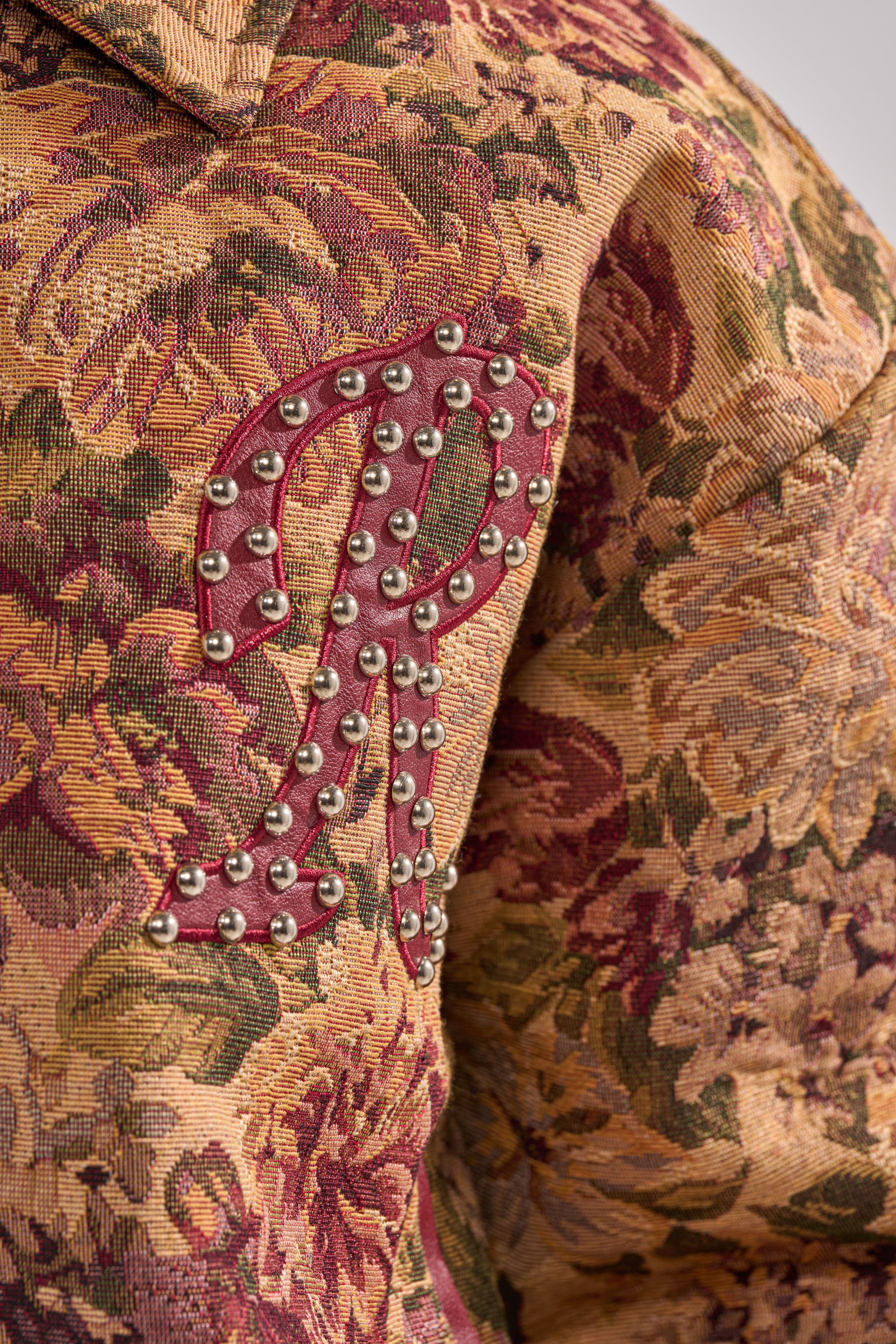 The SLIDE ON ME TAPESTRY BOMBER features a close-up of its floral jacquard design, highlighted by a large red "R" patch with silver studs on the shoulder. 11