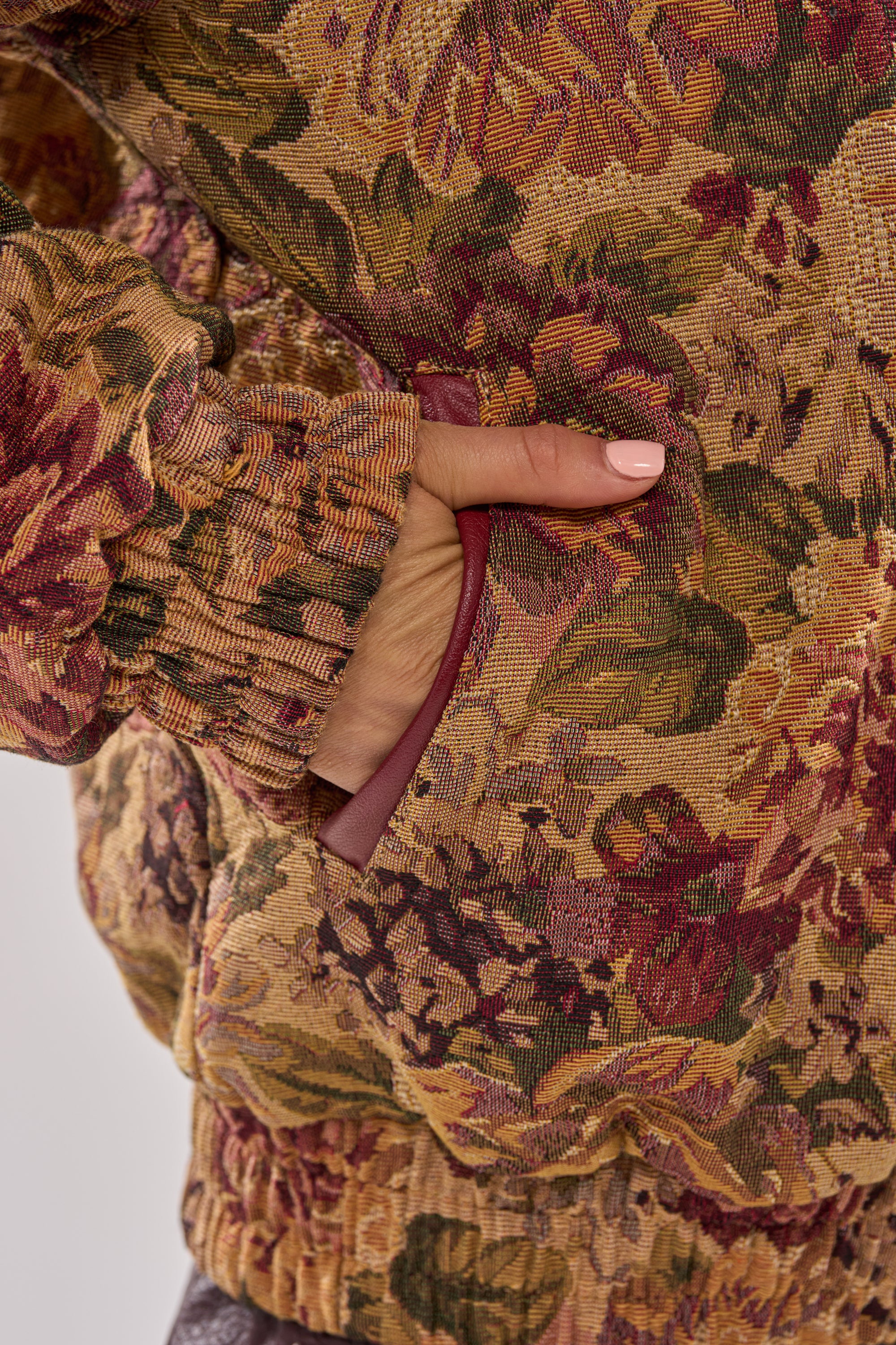 A close-up of a hand with light pink nail polish in the pocket of the SLIDE ON ME TAPESTRY BOMBER, featuring rich brown, gold, red, and green jacquard floral tapestry fabric. 12