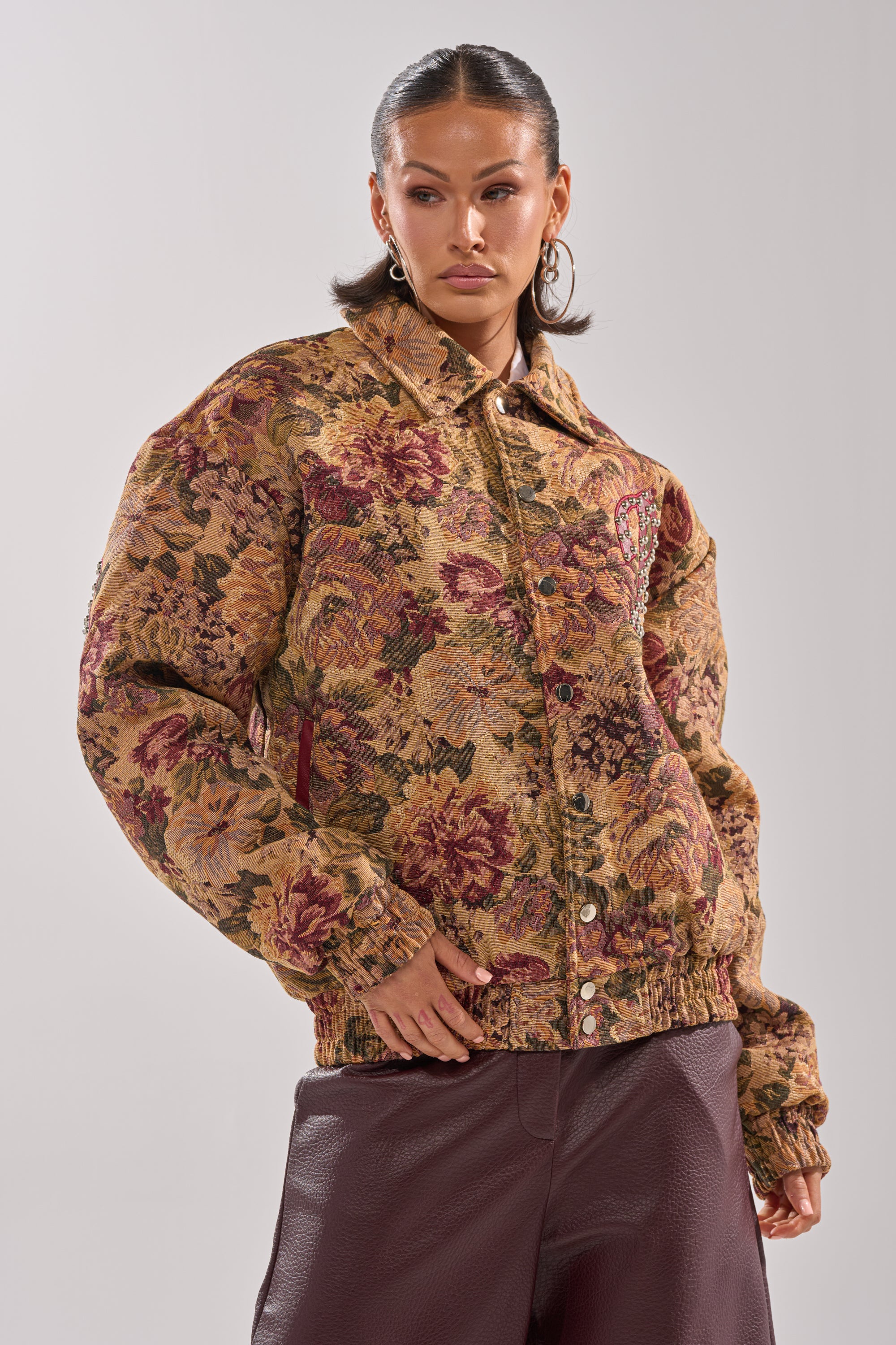 A woman in the SLIDE ON ME TAPESTRY BOMBER jacket with floral print and large buttons, paired with burgundy leather pants, stands against a plain light background, looking confidently to the side. 1