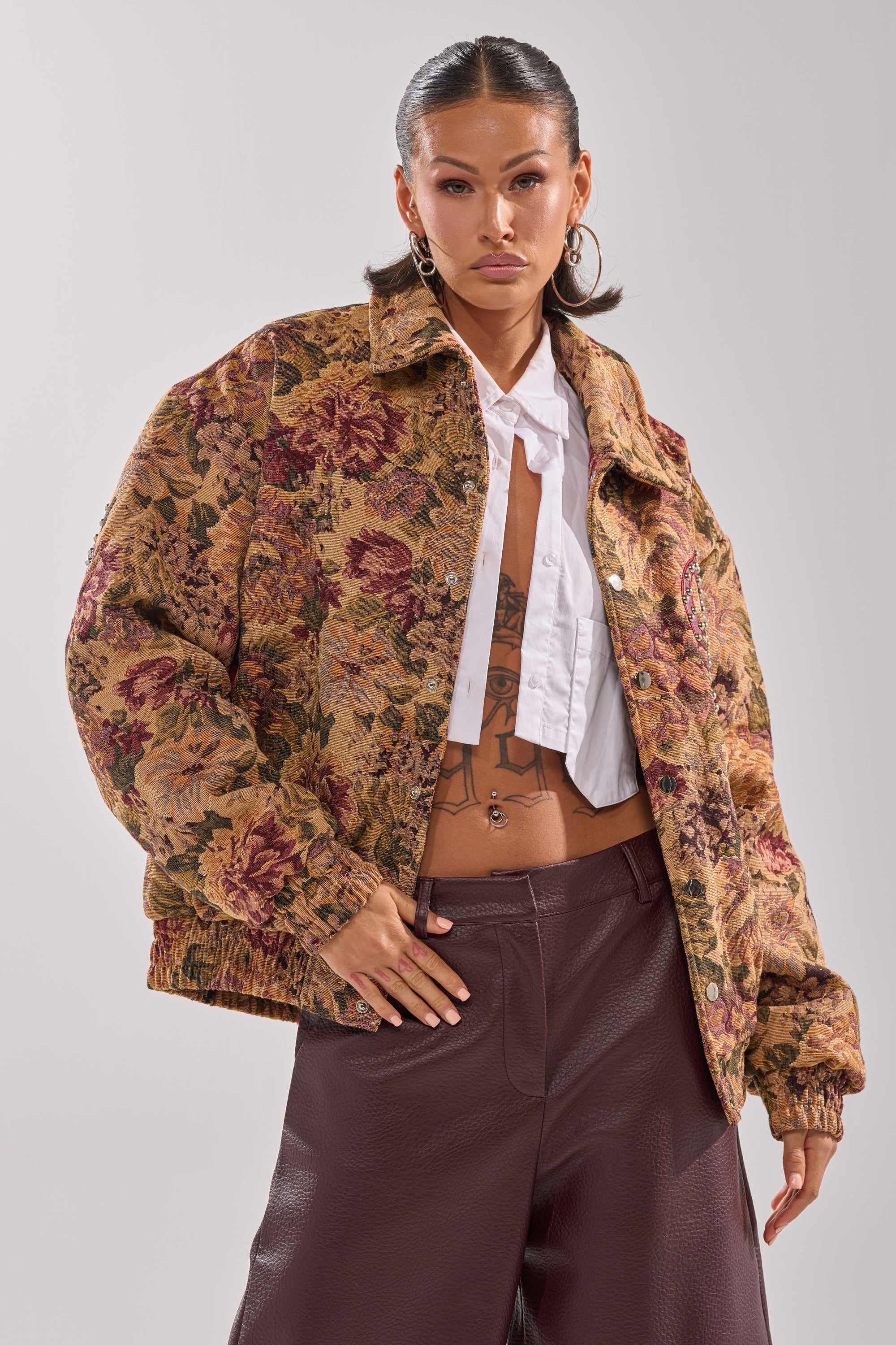 A woman models the SLIDE ON ME TAPESTRY BOMBER with a floral print, open over a white shirt, paired with high-waisted brown pants. She has slicked-back hair, large hoop earrings, and poses neutrally against a plain background. 2