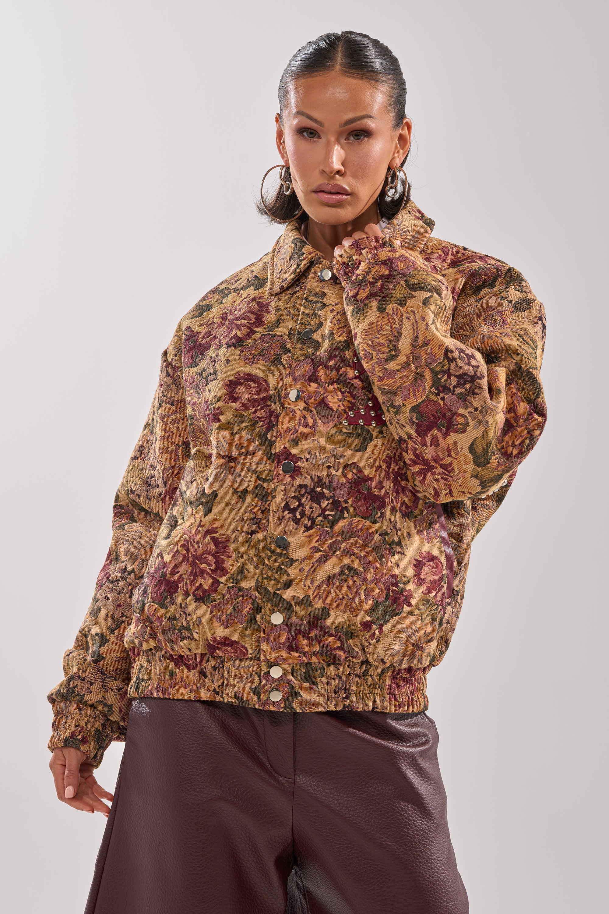 A woman poses confidently against a plain light background wearing the SLIDE ON ME TAPESTRY BOMBER with floral print and large buttons, paired with burgundy leather pants. 4