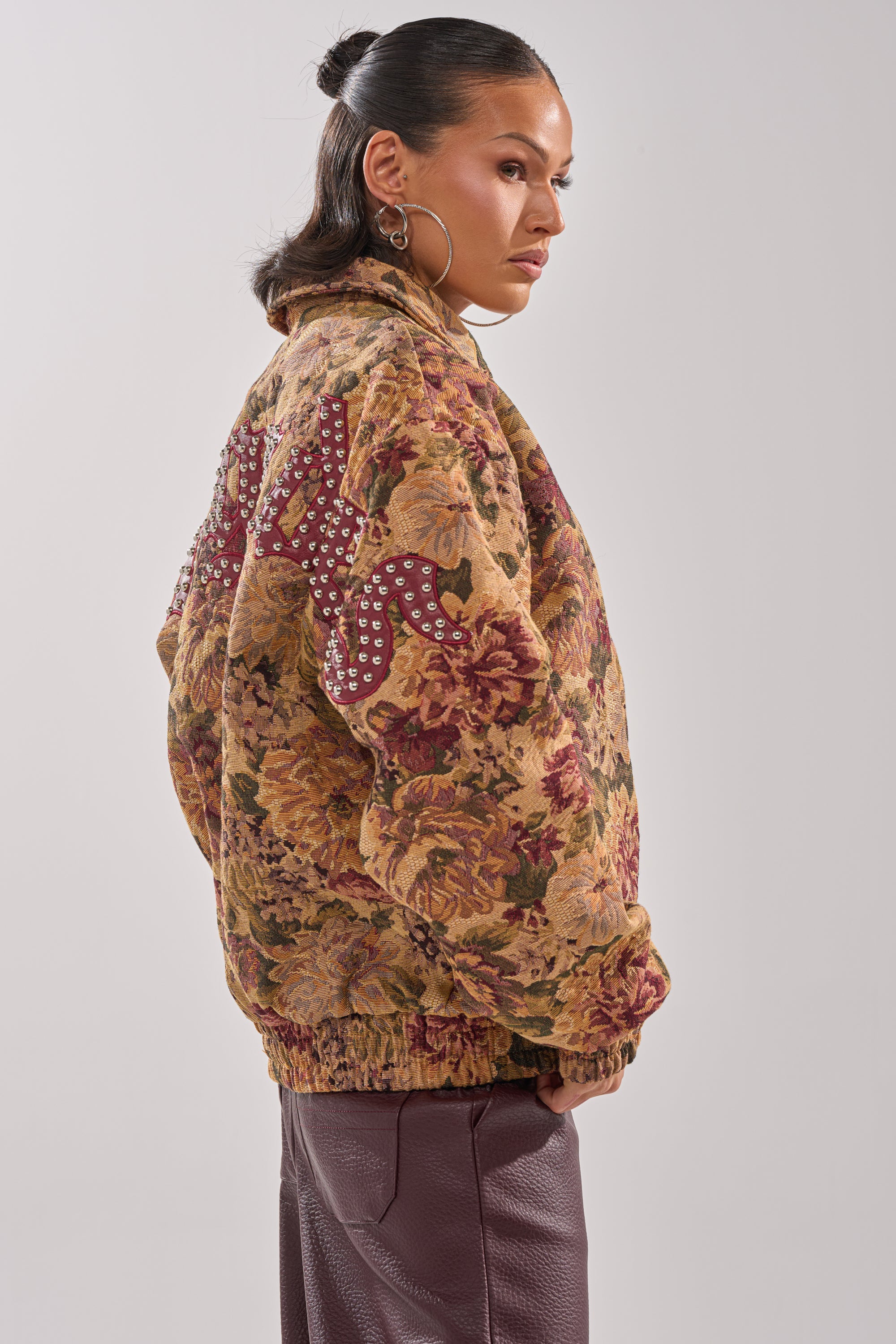 A woman stands sideways, looking serious in the SLIDE ON ME TAPESTRY BOMBER with studded back text and textured maroon pants. She wears hoop earrings and her sleek dark hair is pulled back. 5