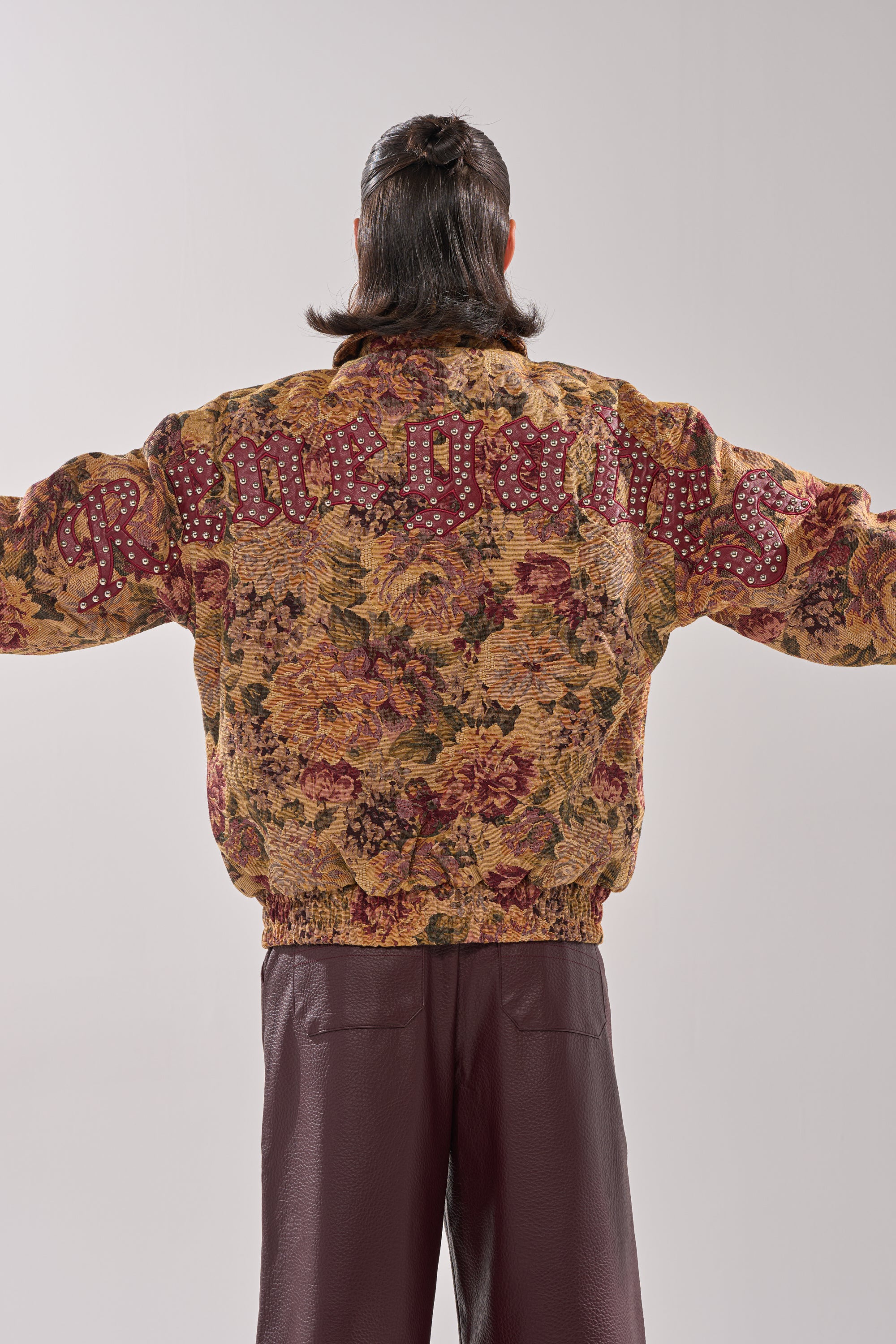 A person with long dark hair stands with arms outstretched, seen from the back, wearing an oversized SLIDE ON ME TAPESTRY BOMBER featuring a floral print and "Rineguns" text, paired with maroon textured pants. 6