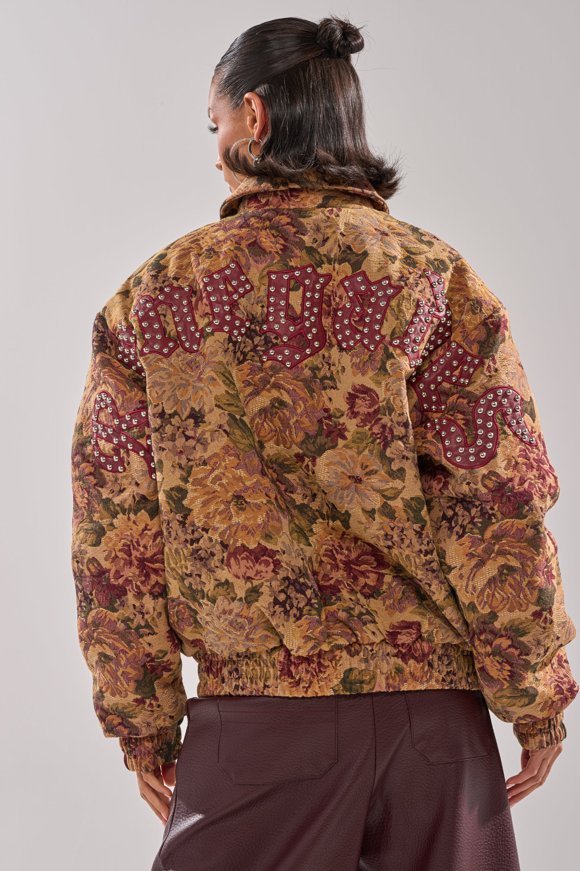 A person with dark hair in a low bun models the SLIDE ON ME TAPESTRY BOMBER, featuring floral jacquard and maroon text with silver studs on the back, paired with textured maroon pants, facing away against a plain background. 7