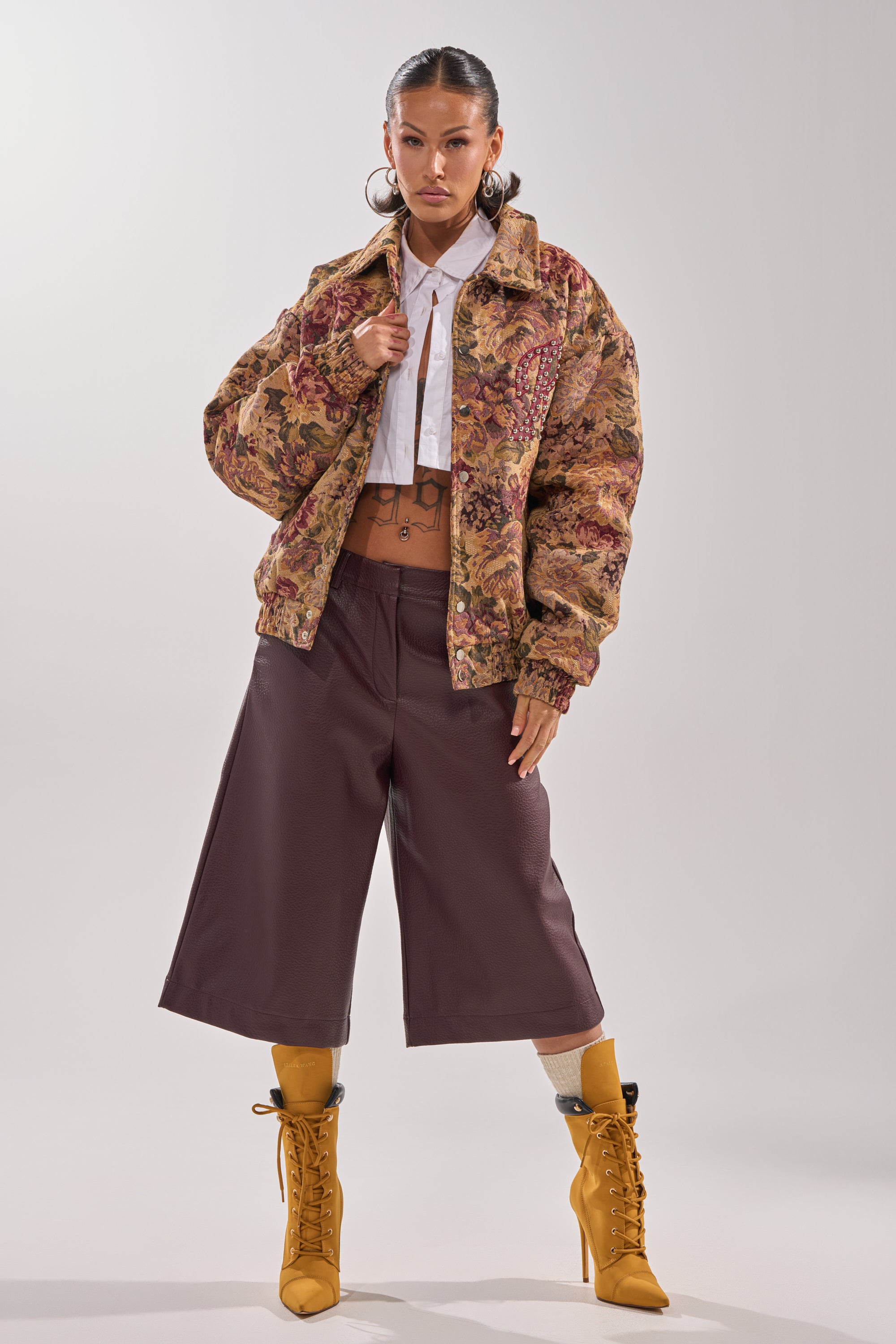A woman stands confidently in the SLIDE ON ME TAPESTRY BOMBER, a white cropped shirt, wide-leg brown culottes, and mustard yellow lace-up boots against a plain light background. 8