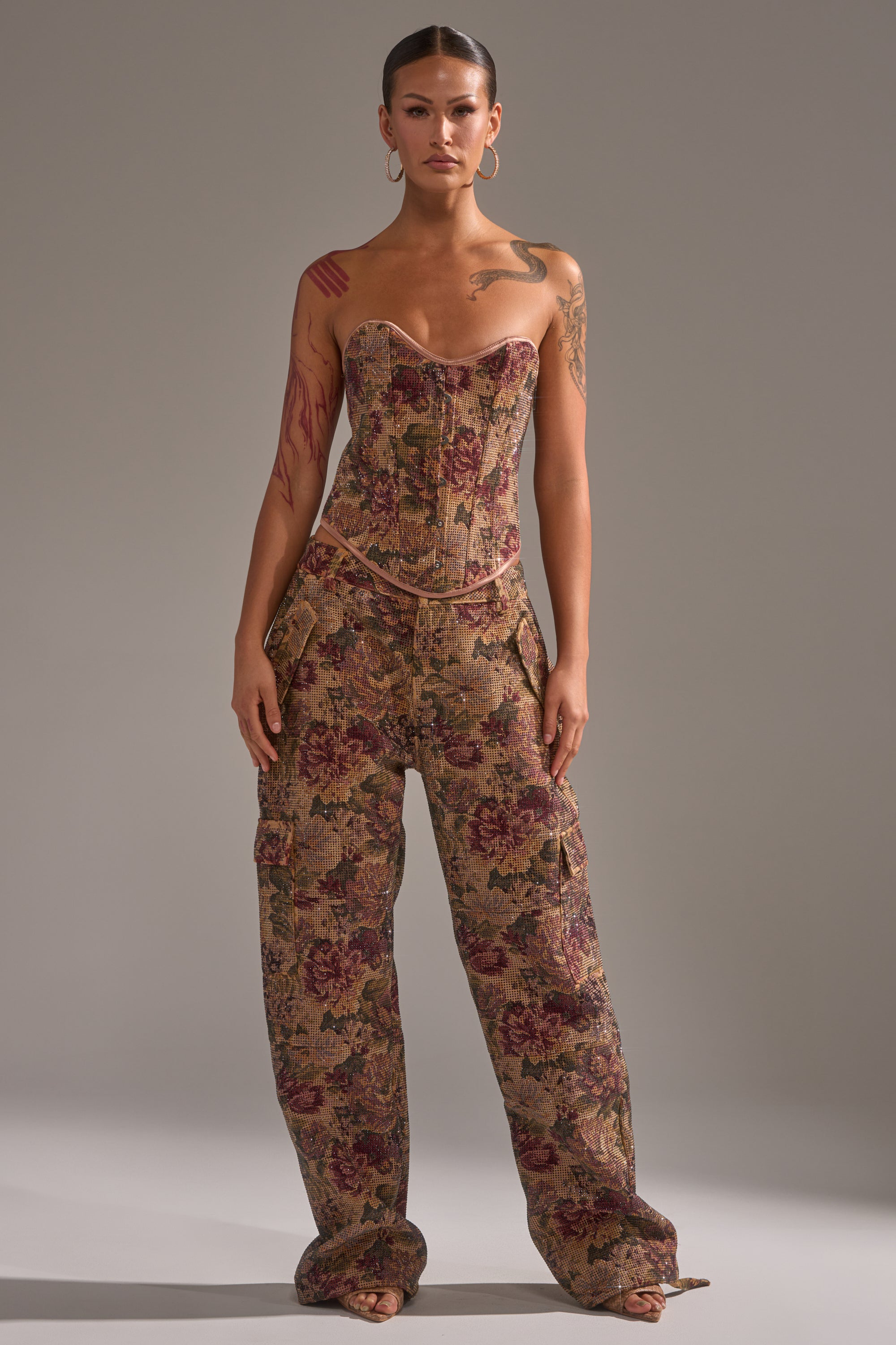 A person faces forward in the GLAM SQUAD RHINESTONE BROCADE TROUSER and a matching floral strapless corset top. They have arm tattoos, hoop earrings, slicked-back hair, and stand against a plain neutral background. 0