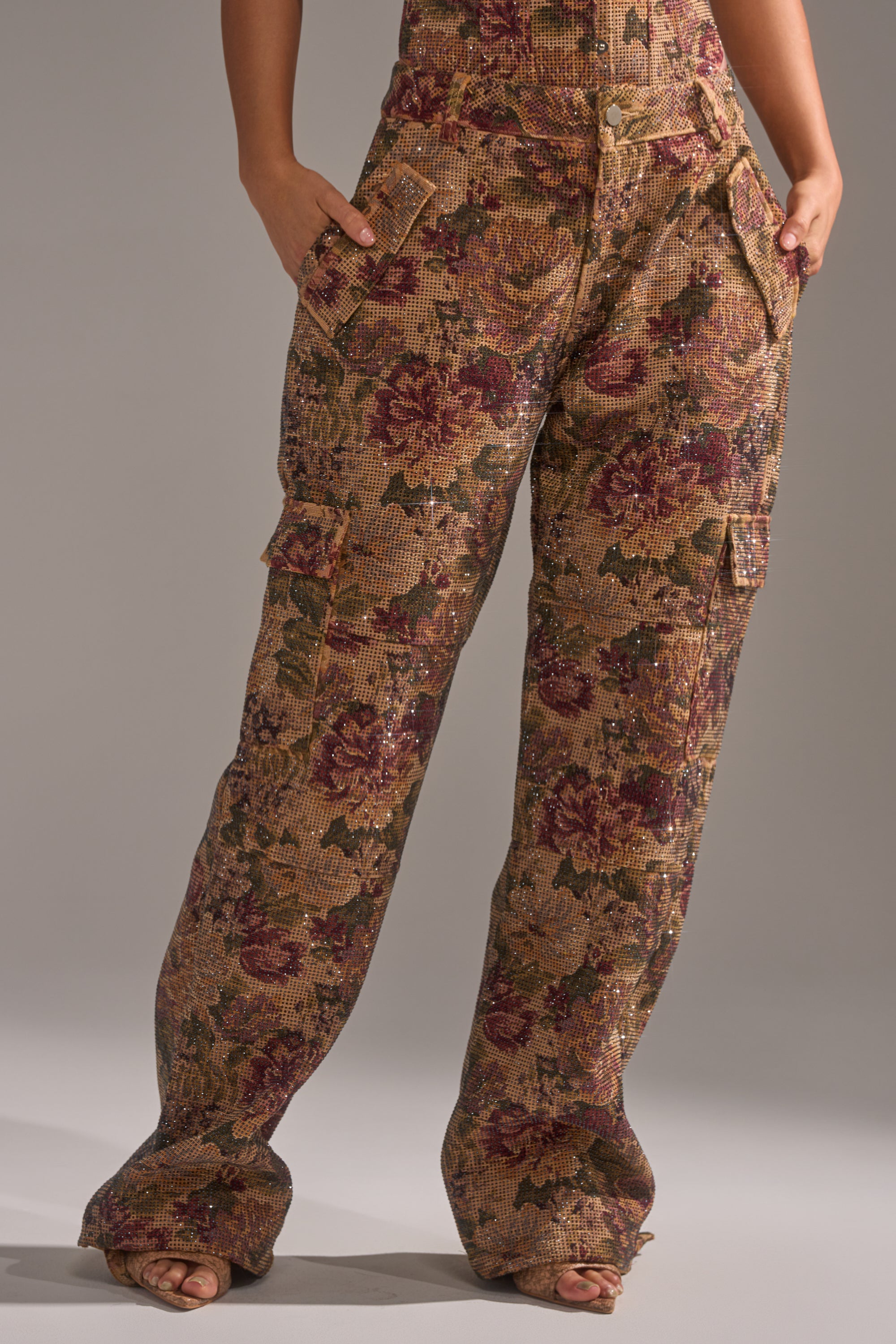 A person wearing the GLAM SQUAD RHINESTONE BROCADE TROUSER—high-waisted, wide-leg cargo pants with a brown, green, and burgundy floral tapestry pattern—stands barefoot with hands in pockets. 1