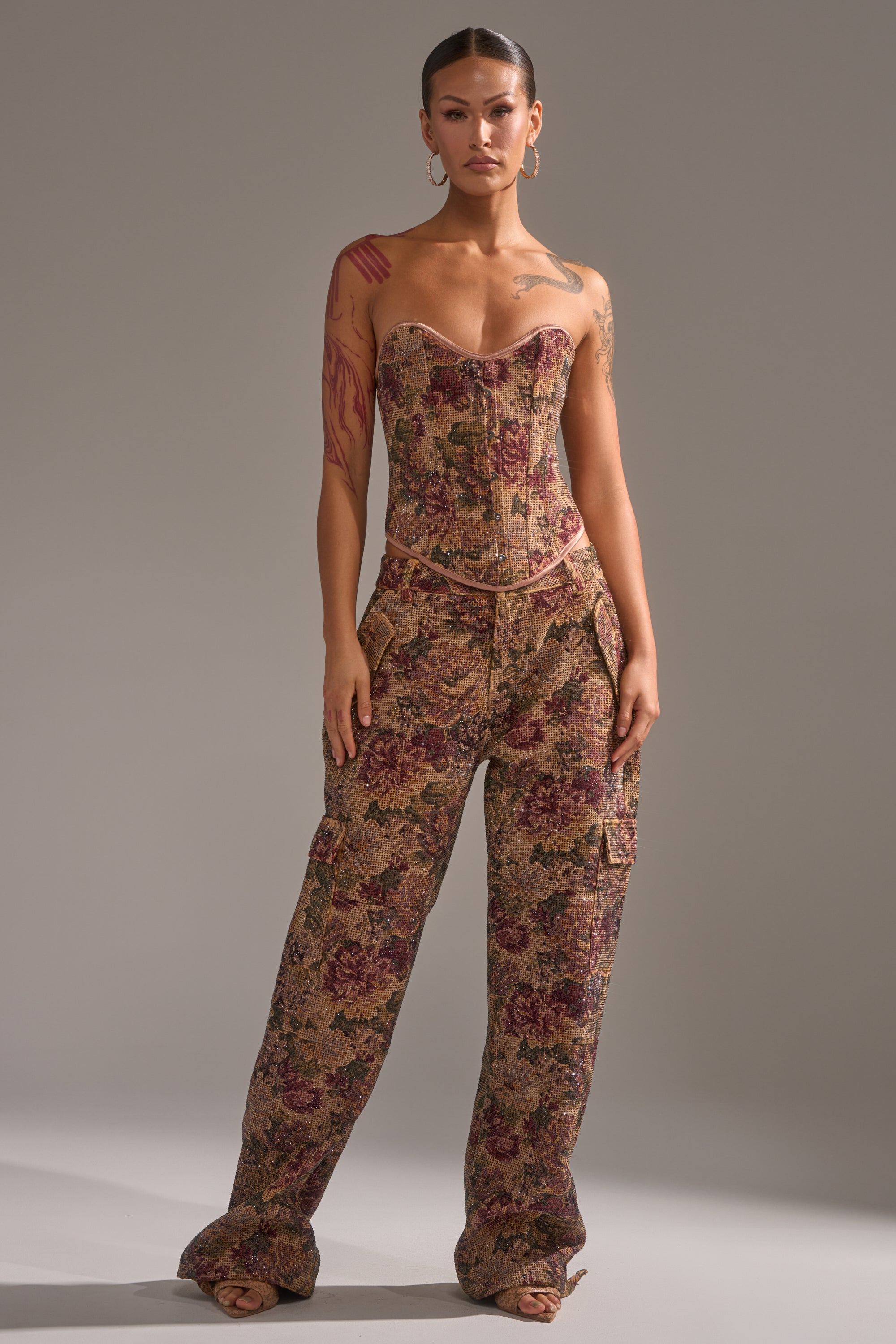 A woman with tattoos on her arms and chest poses against a plain gray background, wearing the GLAM SQUAD RHINESTONE BROCADE TROUSER—loose, floral jacquard pants in a brown pattern paired with a fitted, strapless corset top. 6