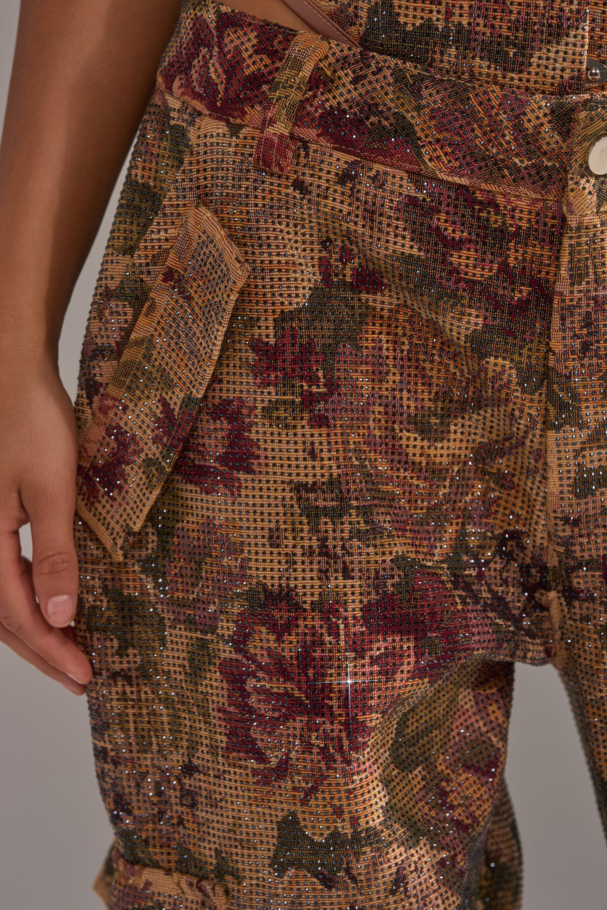 A close-up of a person's lower torso and hand wearing the GLAM SQUAD RHINESTONE BROCADE TROUSER, showcasing brown, red, and green floral jacquard with a glittery finish, flap pocket, and visible white button. 7