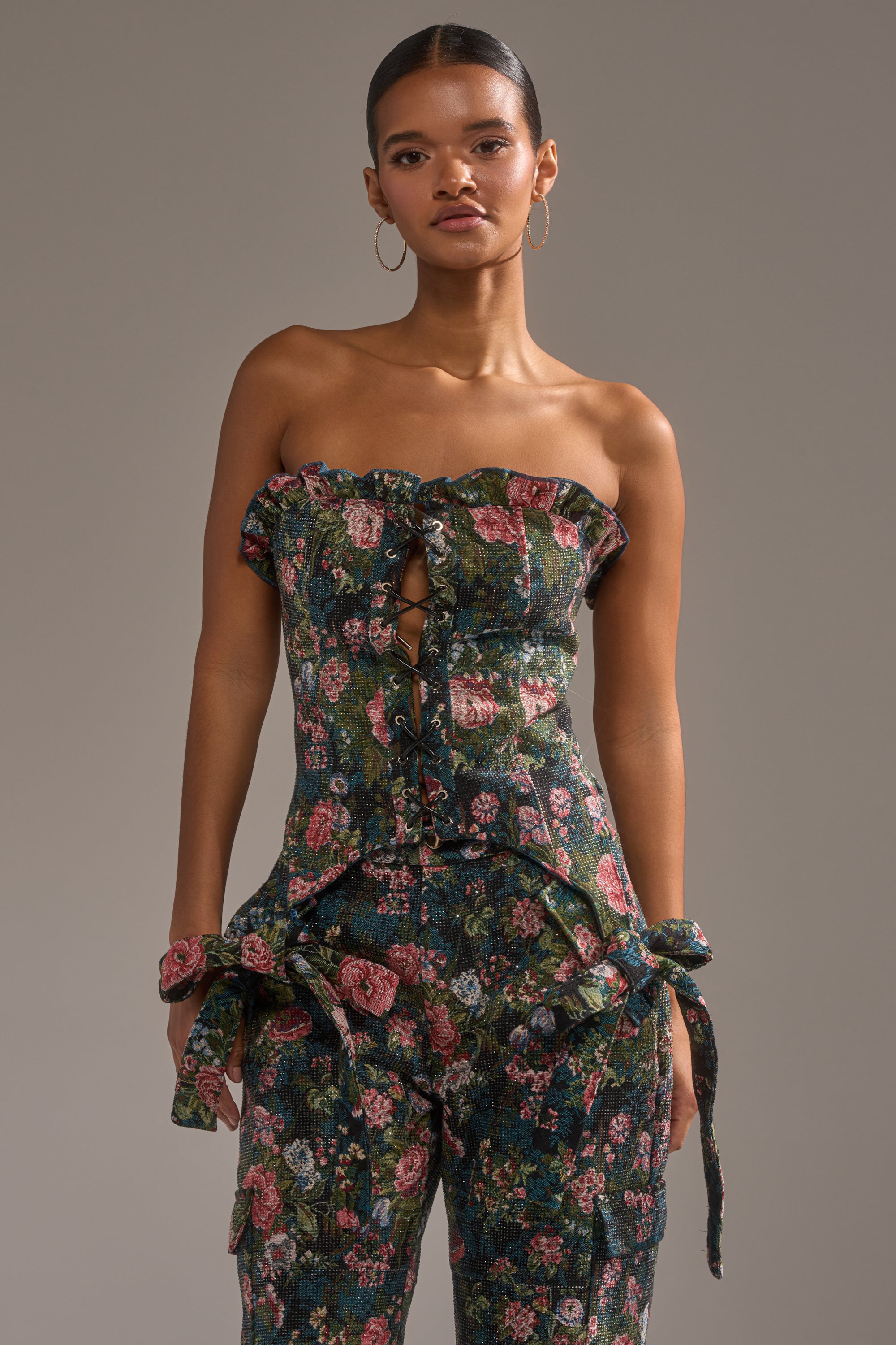A woman wears the I WANT YOU FLORAL RHINESTONE CORSET, a strapless floral jumpsuit with a lace-up front and tied bows at the hips. She accessorizes with hoop earrings and has her hair pulled back, posing against a plain background. 0