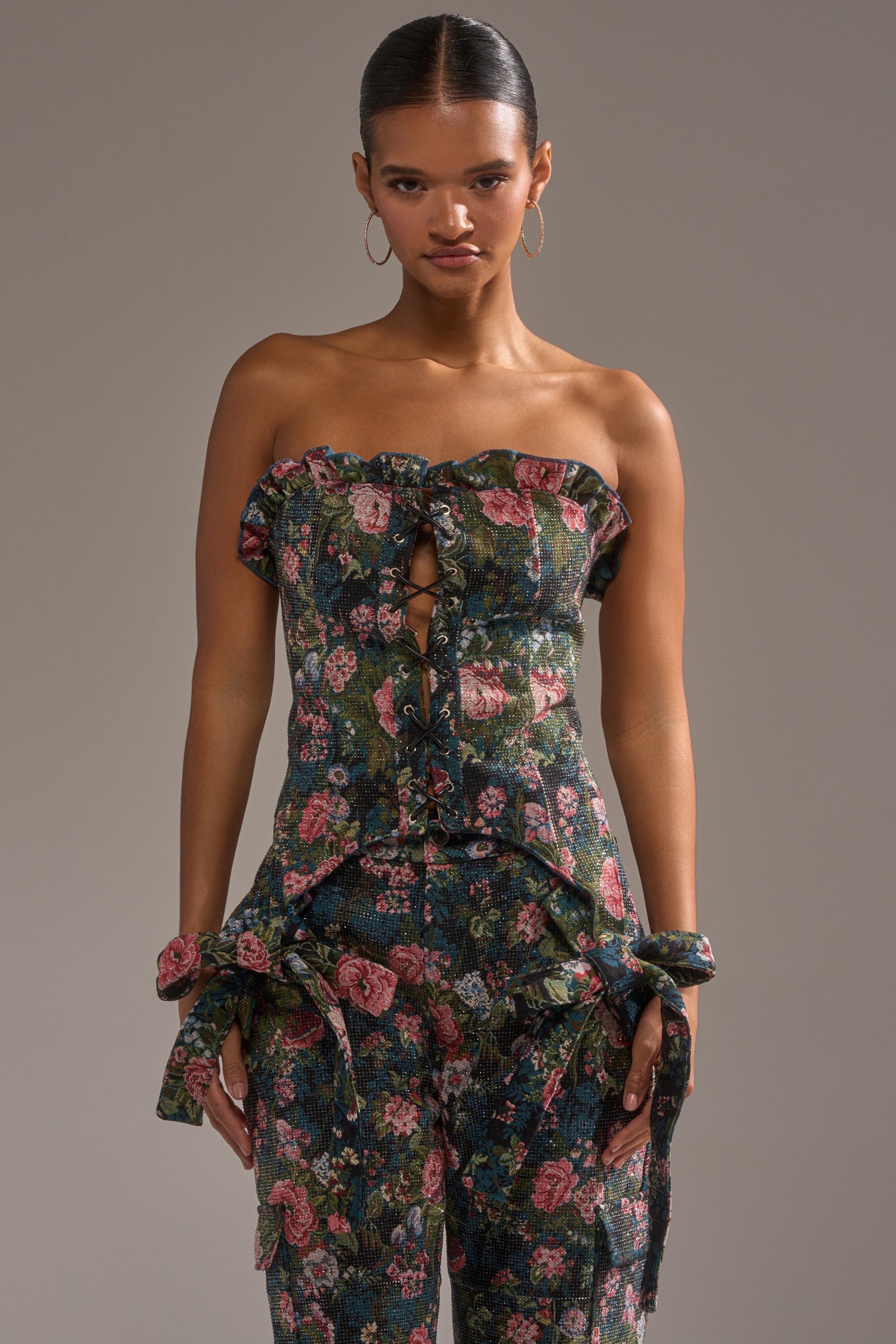 A woman stands confidently against a neutral background, wearing the I WANT YOU FLORAL RHINESTONE CORSET—strapless, vintage-inspired, with a floral pattern, lace-up front, and fabric bows on the hips. 1