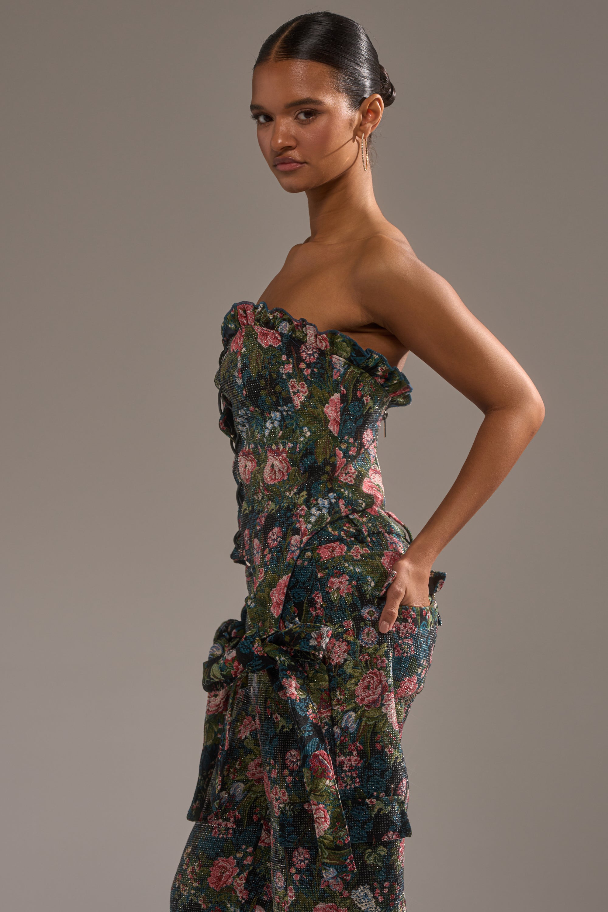A woman wearing the I WANT YOU FLORAL RHINESTONE CORSET dress stands in profile against a gray background, showcasing its strapless floral pattern and structured fit, with one hand on her hip and her hair in a sleek bun. 2