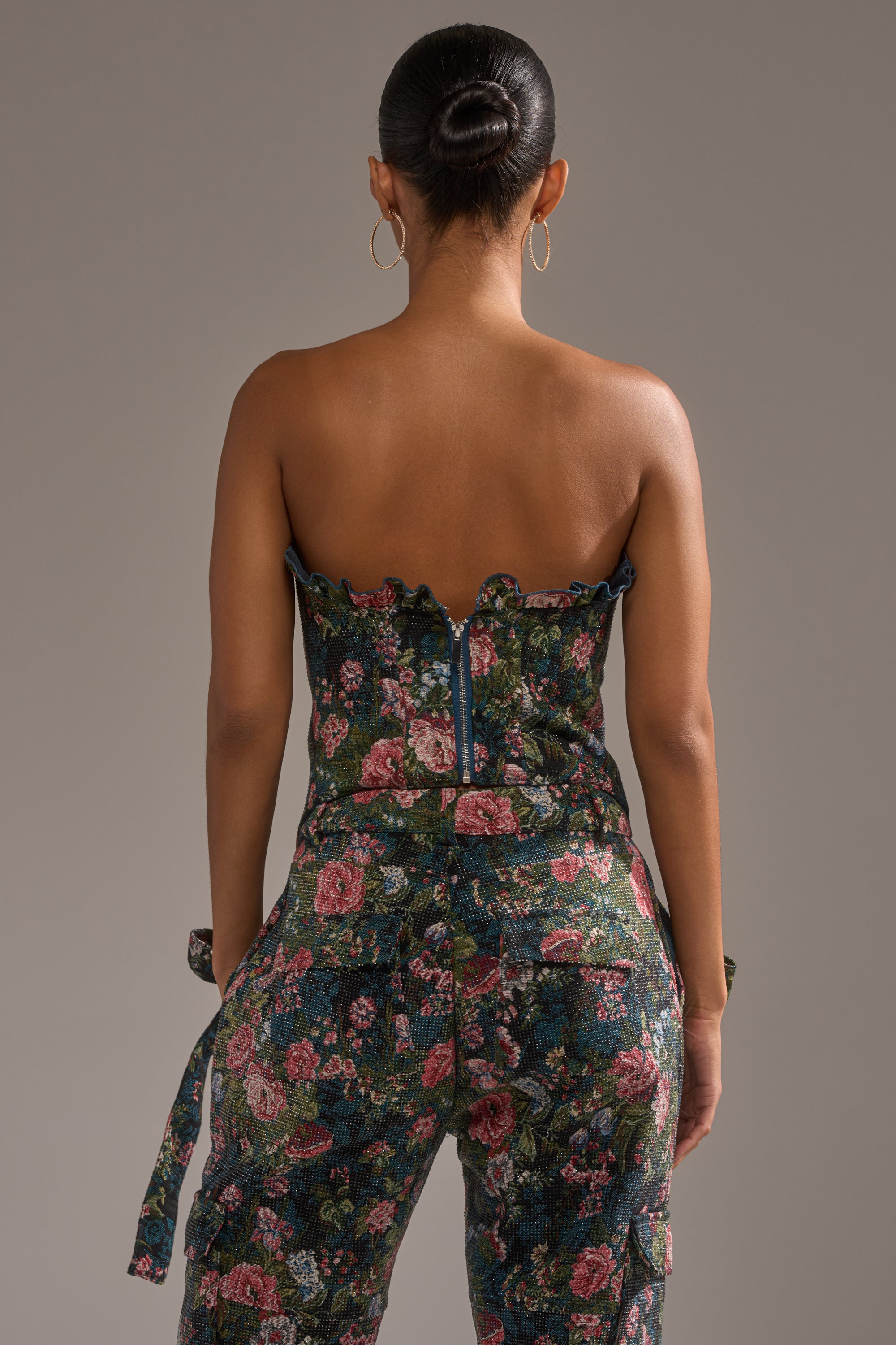 A person with hair in a neat bun is seen from behind wearing the I WANT YOU FLORAL RHINESTONE CORSET top and matching pants in green, pink, and red shades, featuring floral rhinestones and a visible back zipper. 3
