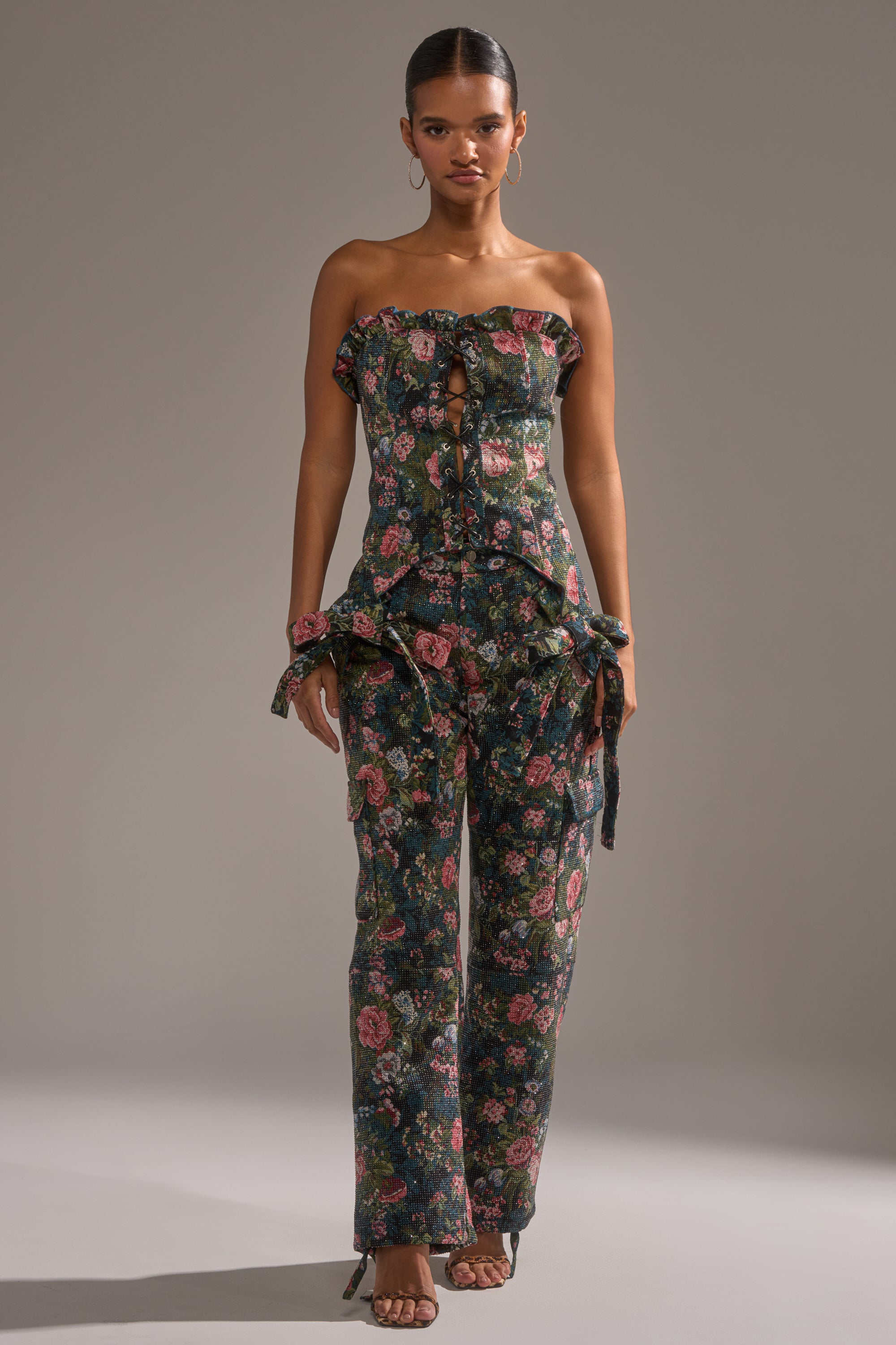 A woman faces forward in the I WANT YOU FLORAL RHINESTONE CORSET, a strapless, ruffled floral piece with a lace-up front evoking vintage corset style. She pairs it with hoop earrings, sandals, and has her hair pulled back against a plain gray backdrop. 5
