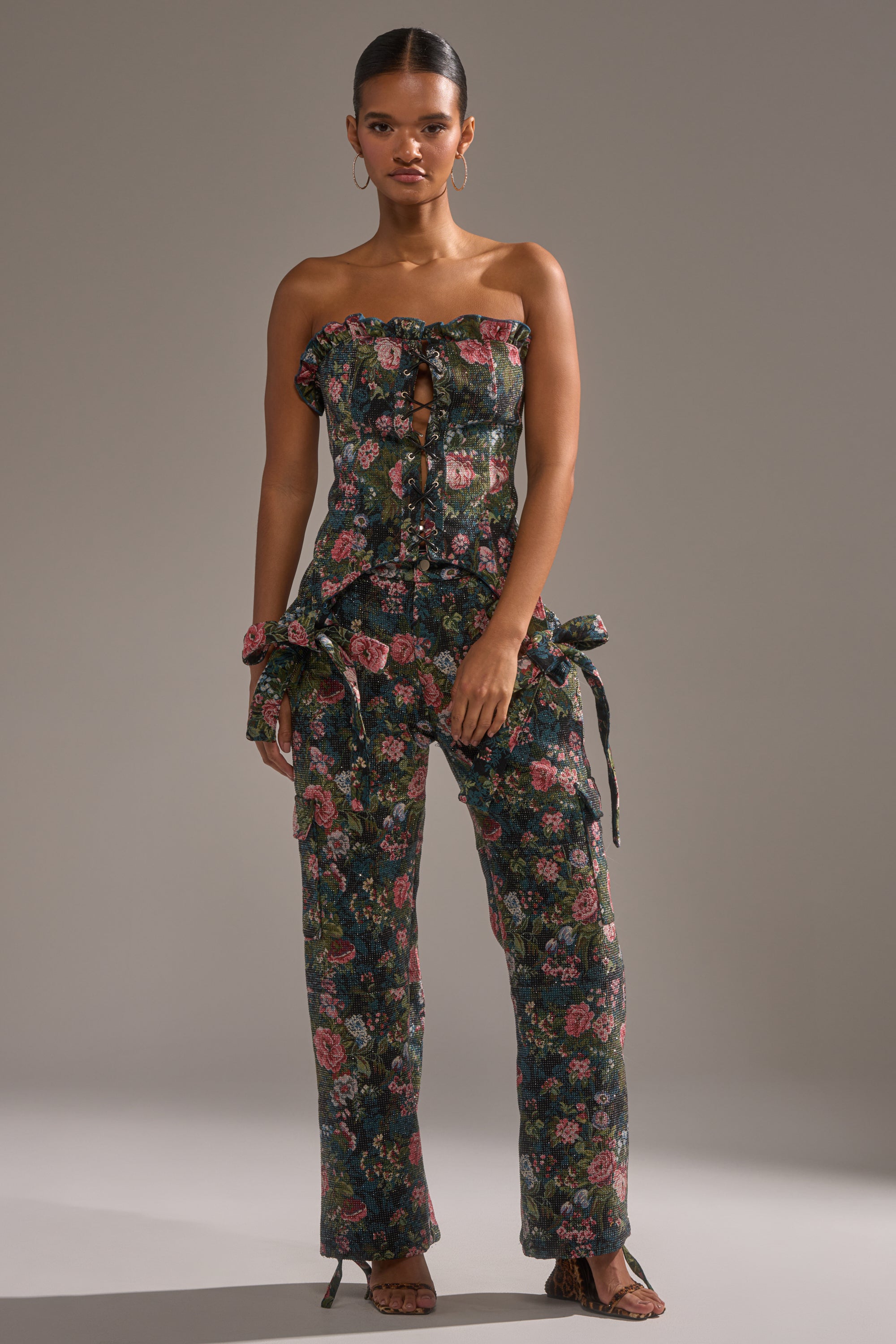 A woman wears the I WANT YOU FLORAL RHINESTONE TROUSER, a dark floral high-waisted pant with rhinestone accents. She pairs it with hoop earrings and slicked-back hair, standing against a plain gray background.
