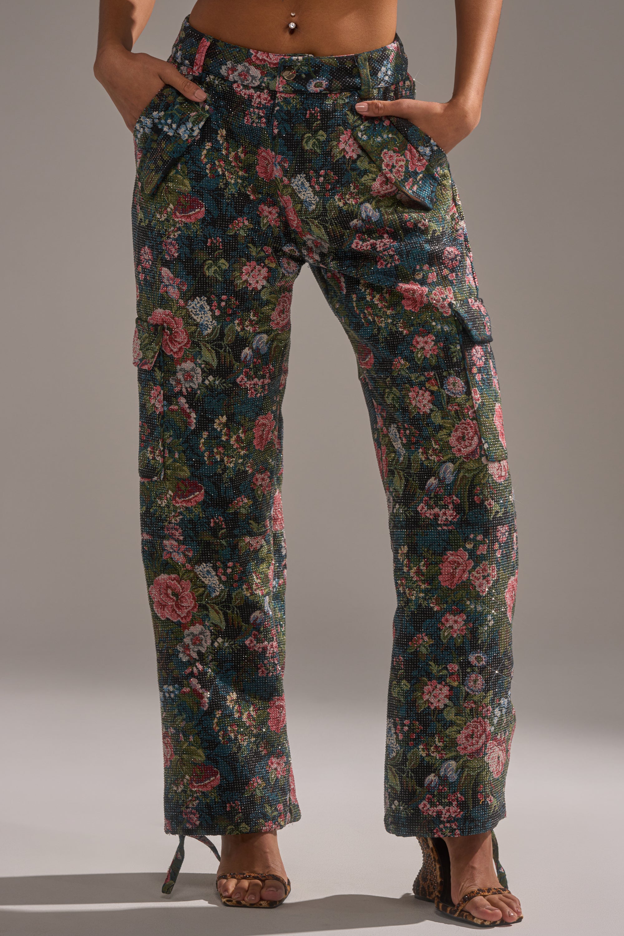 Wearing the I WANT YOU FLORAL RHINESTONE TROUSER—high-waisted, straight-leg pants with a colorful floral rhinestone design and front pockets—she pairs them with open-toe heeled sandals, exposing some midriff.