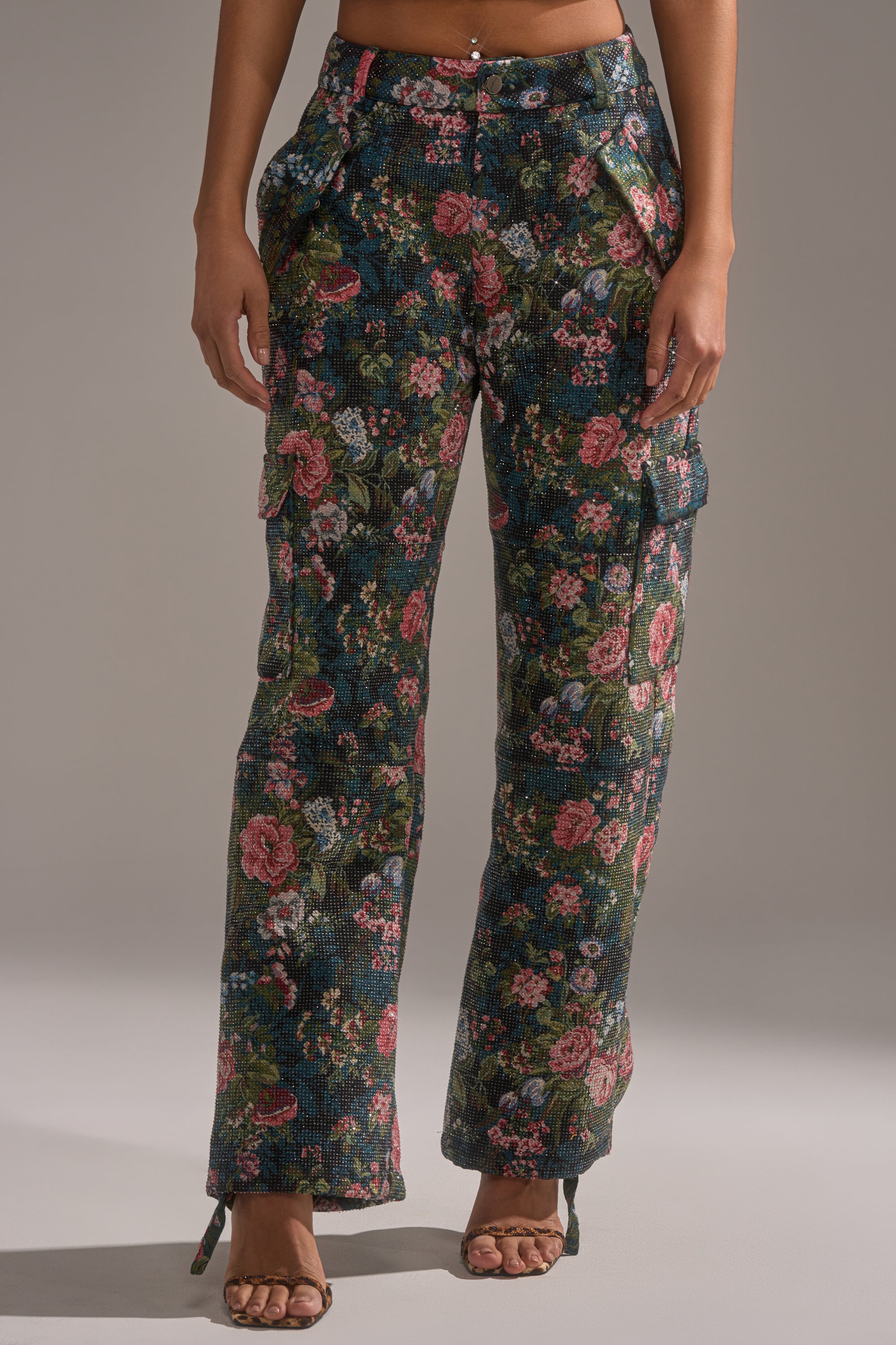 Someone wears the I WANT YOU FLORAL RHINESTONE TROUSER—high-waisted, dark floral pants with rhinestone details and side pockets—paired with strappy high-heeled sandals, standing against a plain background.