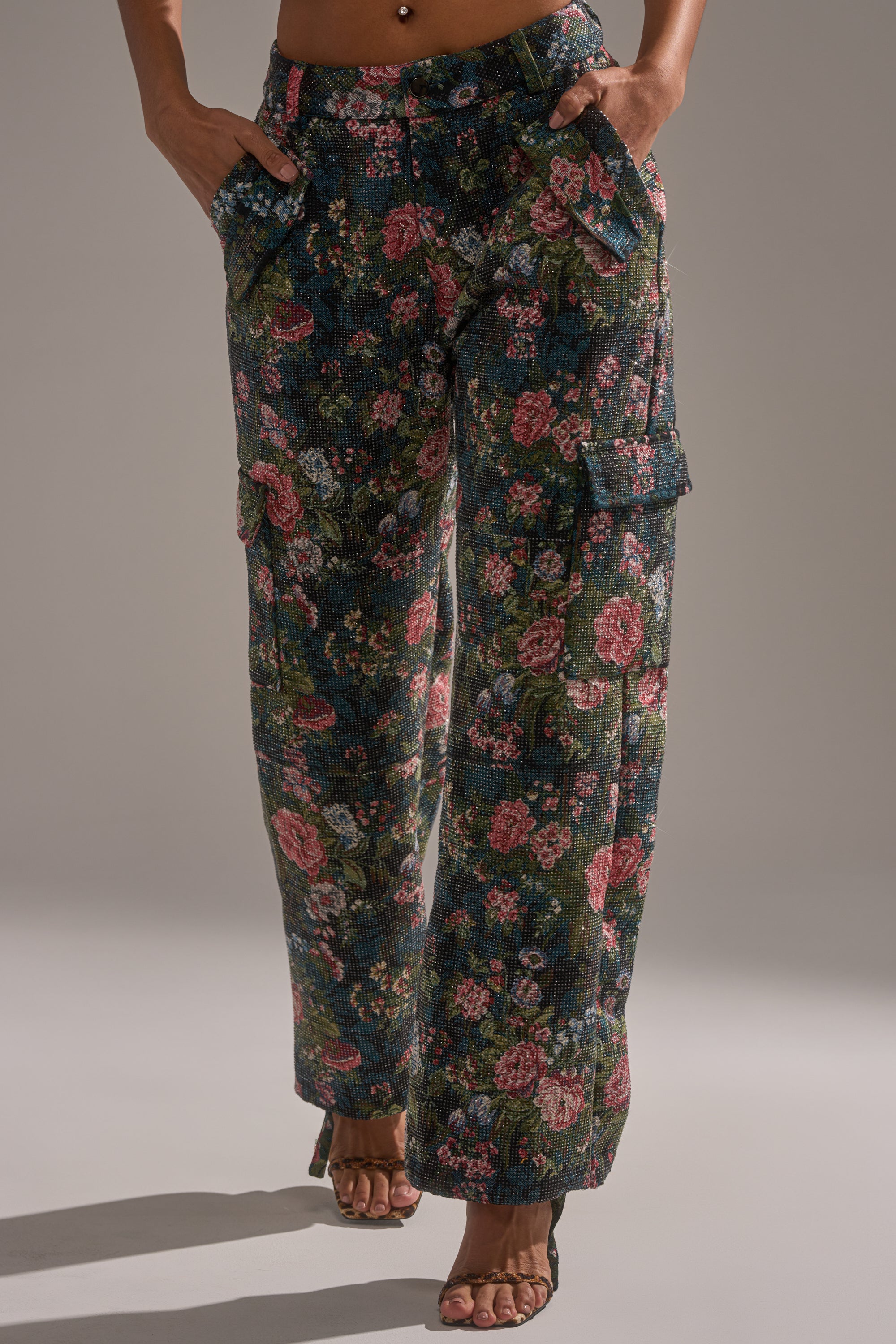 A person stands against a neutral background, hands in pockets and feet in open-toe heels, wearing the I WANT YOU FLORAL RHINESTONE TROUSER—high-waisted, wide-leg cargo pants with a dark floral print and multiple pockets.