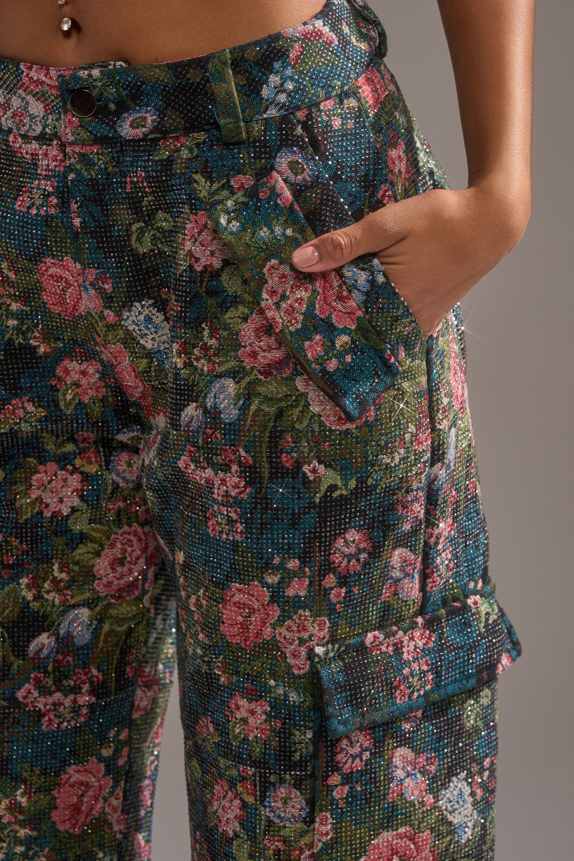 Wearing the I WANT YOU FLORAL RHINESTONE TROUSER—high-waisted with vibrant pink, red, and green patterns—a hand in the pocket and a visible belly button piercing make it ideal for bold statement suiting.