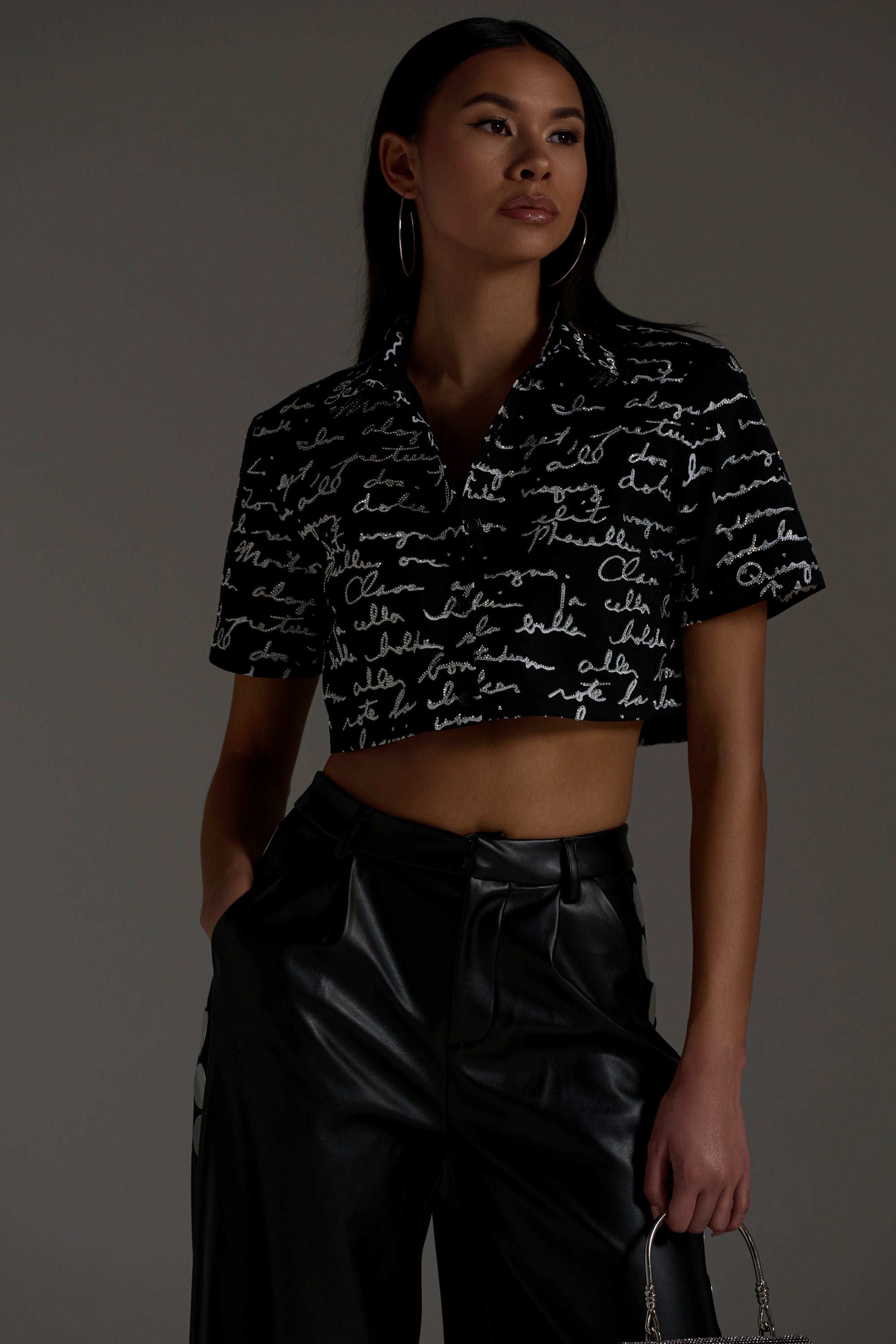 A woman stands confidently in the CHANGES BUTTON DOWN, paired with high-waisted black leather pants and a small handbag against a plain, neutral background.