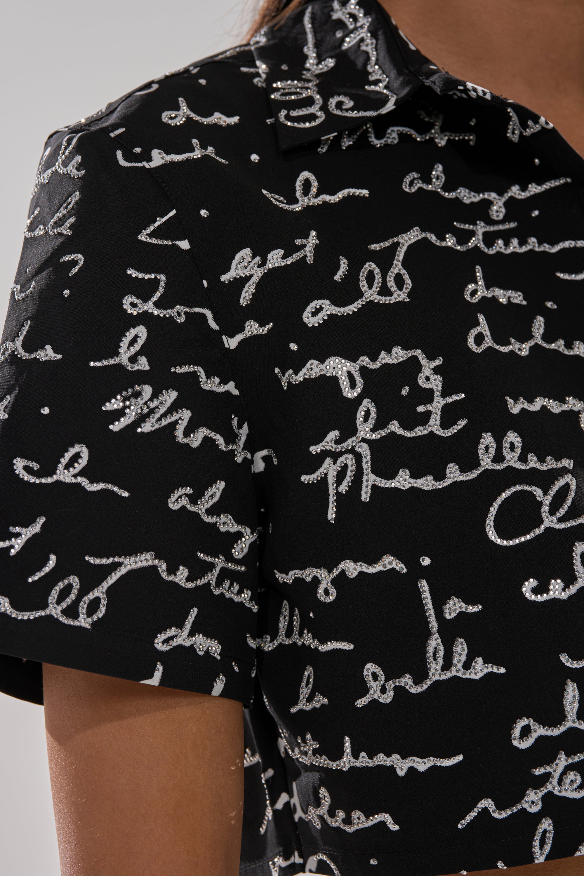 Close-up of the black CHANGES BUTTON DOWN from AKIRA Label, with silver glittery cursive patterns, a collar, and modeled by a person with medium-brown skin.
