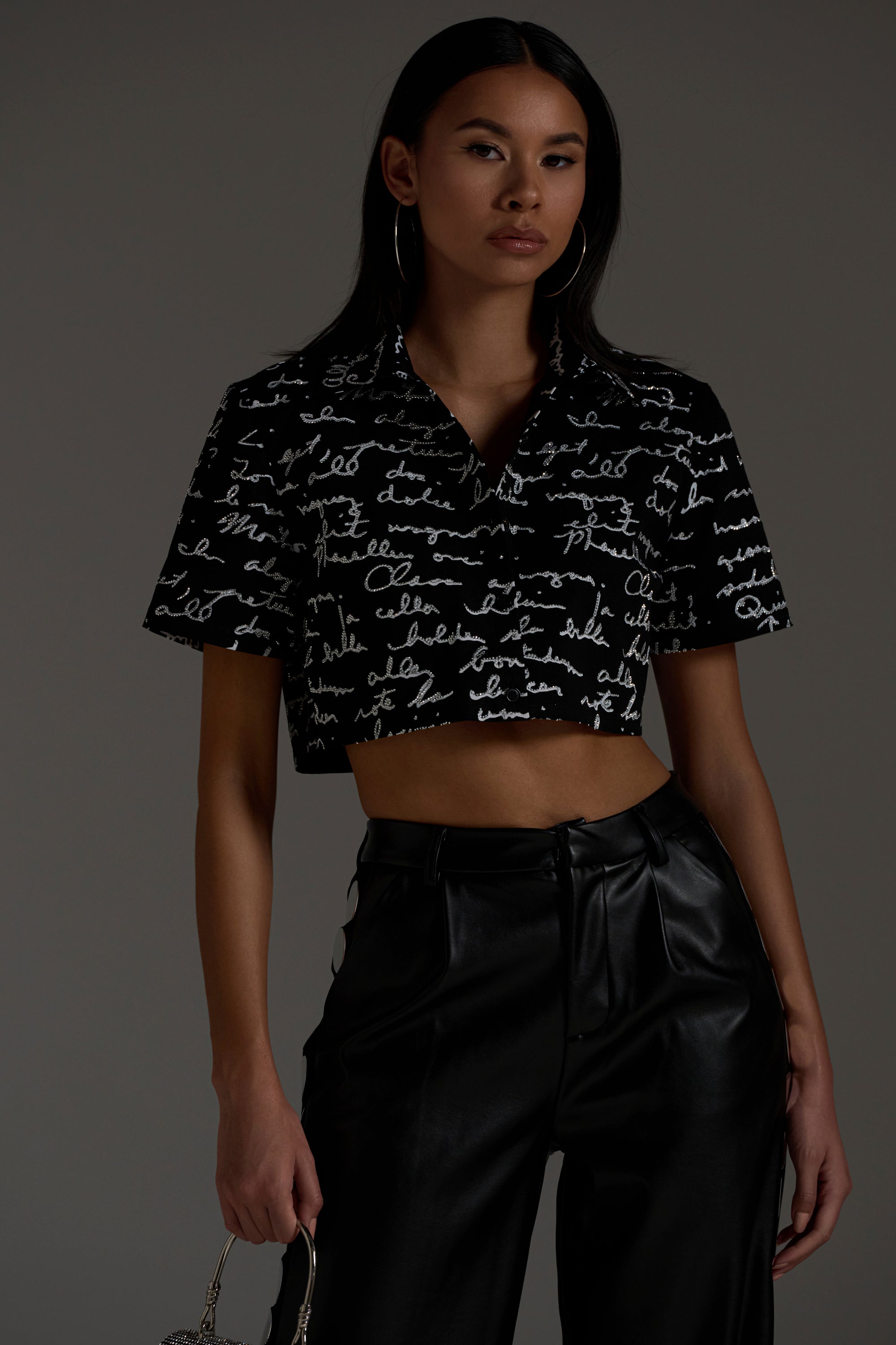 A woman wearing the CHANGES BUTTON DOWN, paired with high-waisted black leather pants, stands against a plain dark background and holds a small purse by her side.