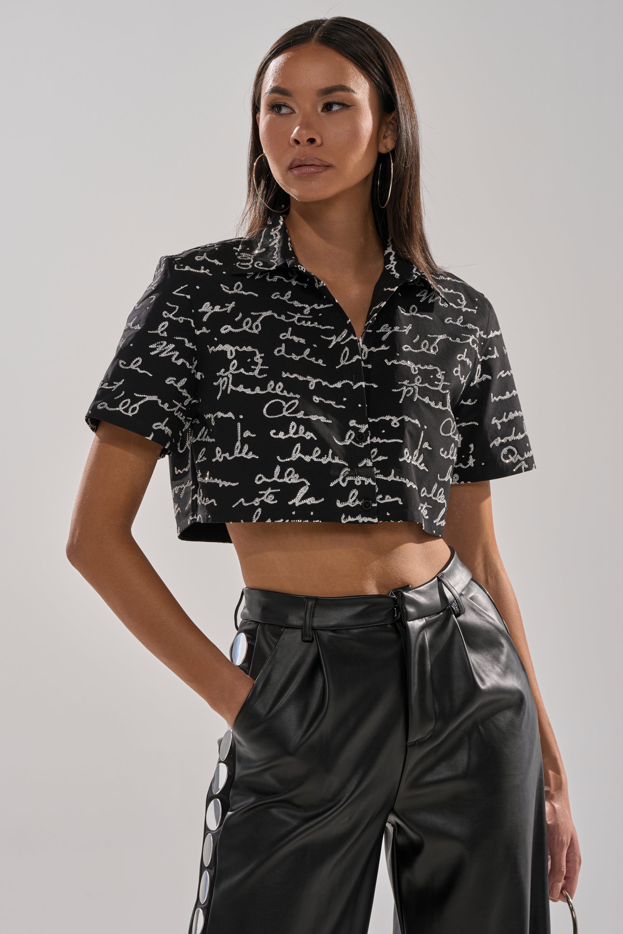 A woman with long straight hair and large hoop earrings stands with one hand in her pocket, wearing the CHANGES BUTTON DOWN featuring white script writing, paired with high-waisted black leather pants with side buttons.