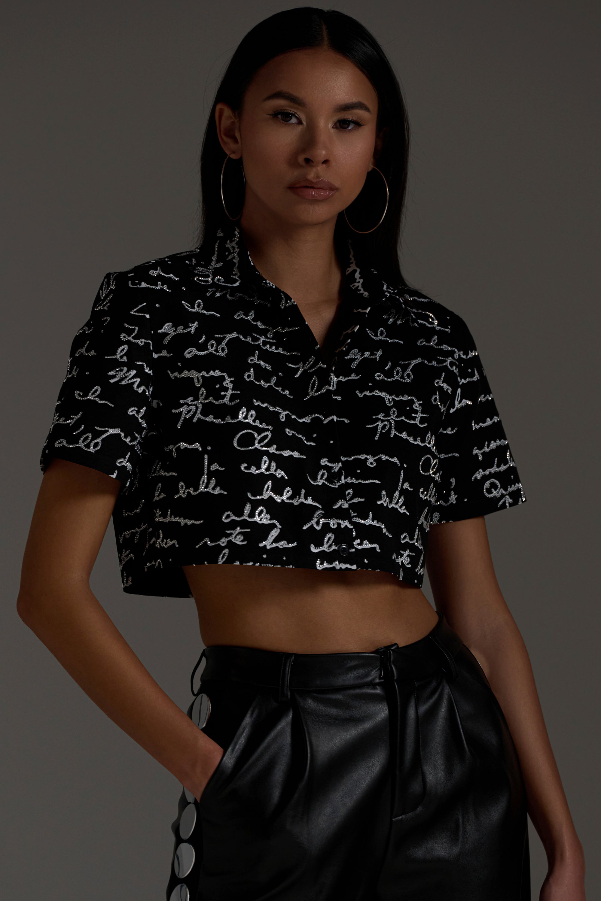 A woman with long dark hair and hoop earrings stands confidently against a dark background, wearing the CHANGES BUTTON DOWN shirt and high-waisted black leather pants.